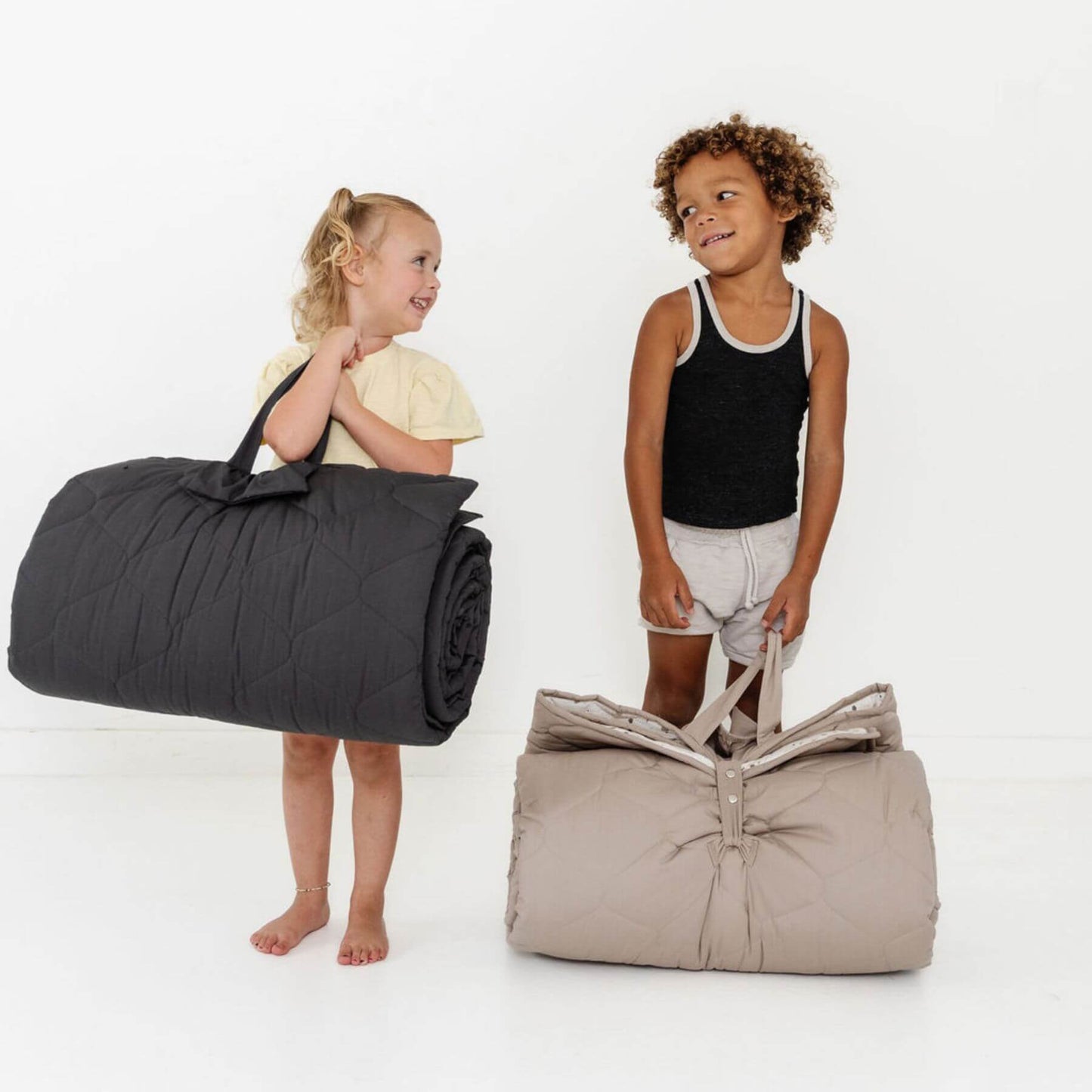 Two children holding rolled-up Toki Kids Picnic Blanket against a white background