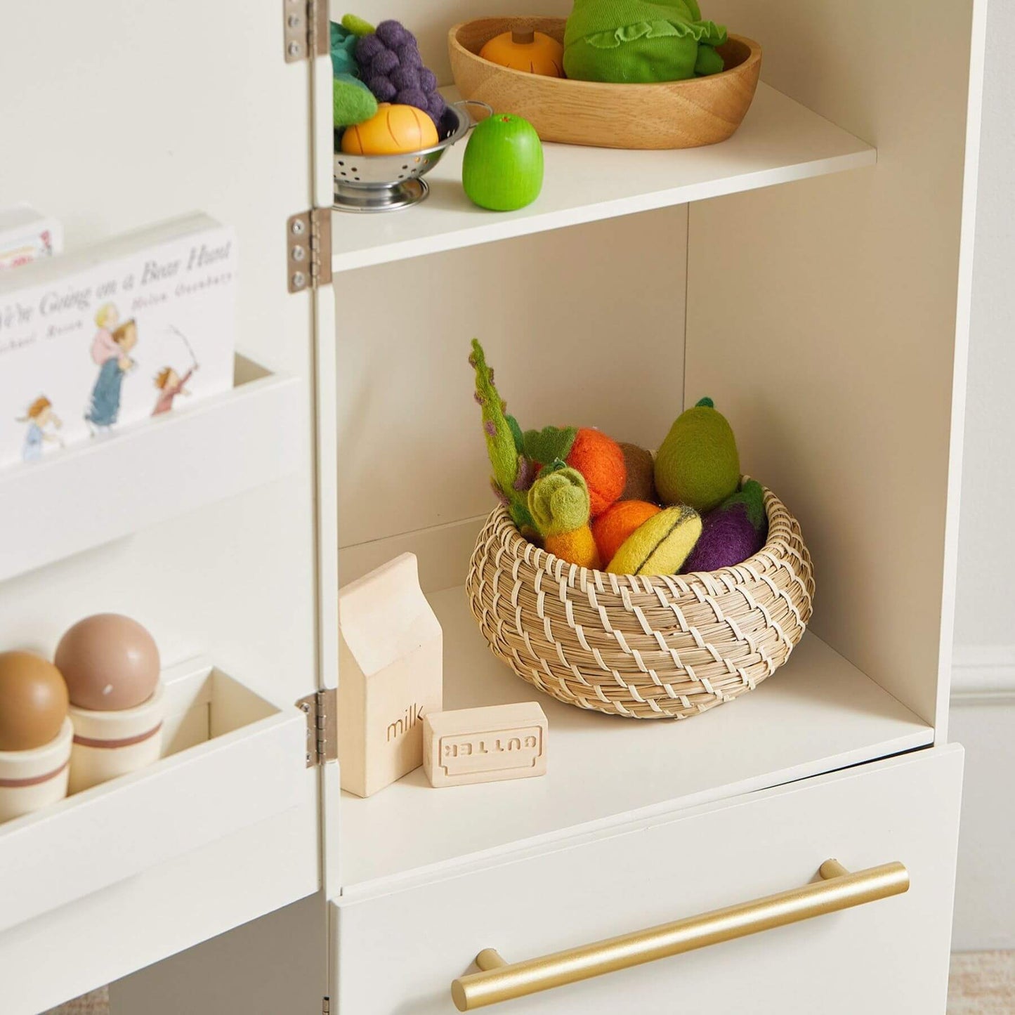Toy fruit and vegetables in a wicker basket on a white shelf with a gold handle.