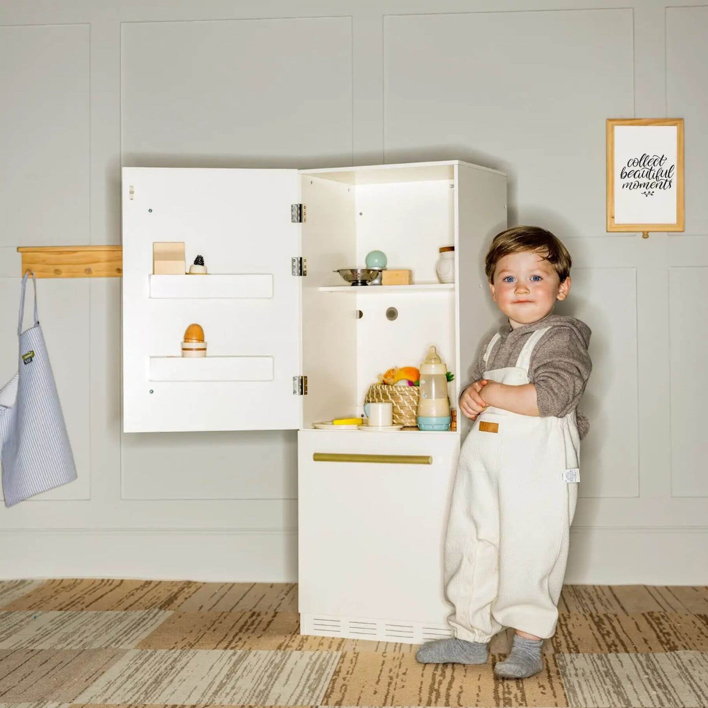 Child standing next to Tiny Land® Cabinet Play Fridge in a room with a framed picture on the wall.