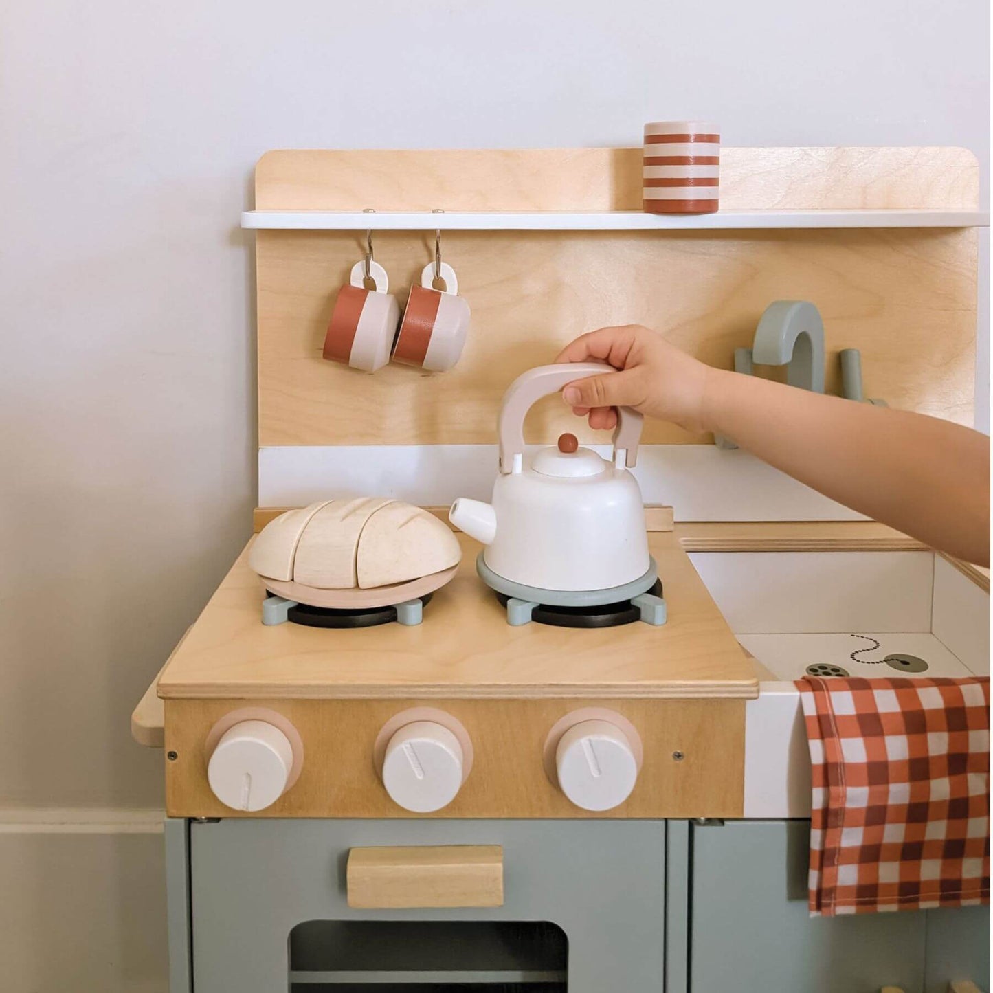 Wooden toy kitchen set with a child's hand placing a white teapot on a stove.