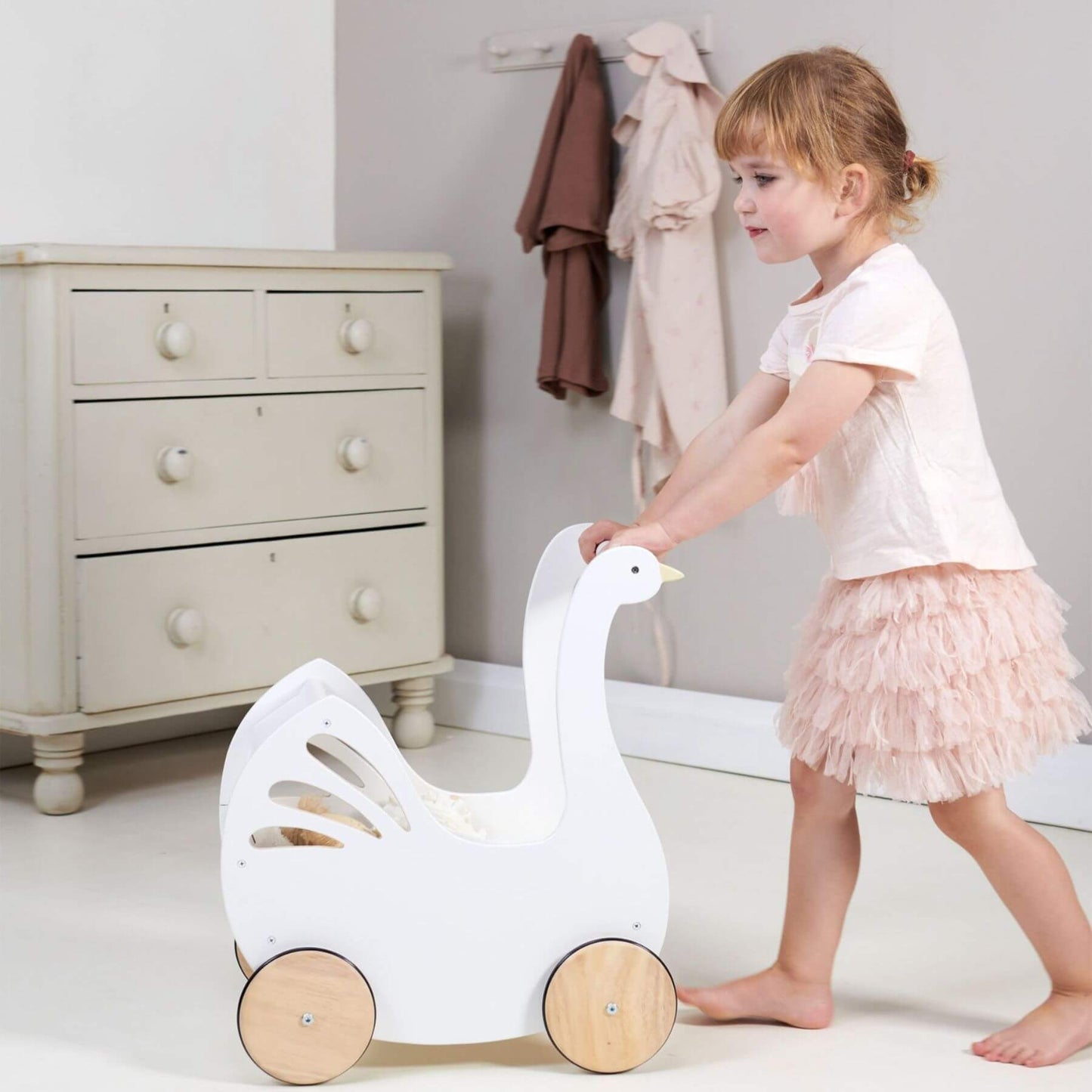 Child playing with Tender Leaf Sweet Swan Pram in a room with a dresser and coats on hooks.