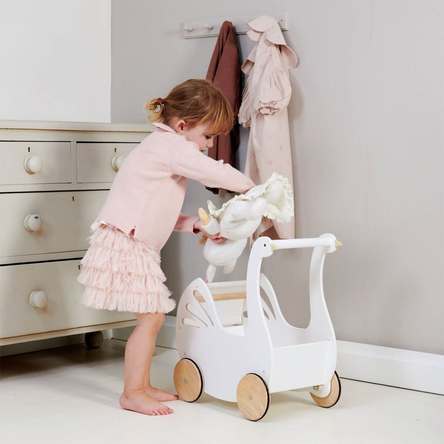 Child playing with Tender Leaf Sweet Swan Pram in a room with white dresser and pink clothing.