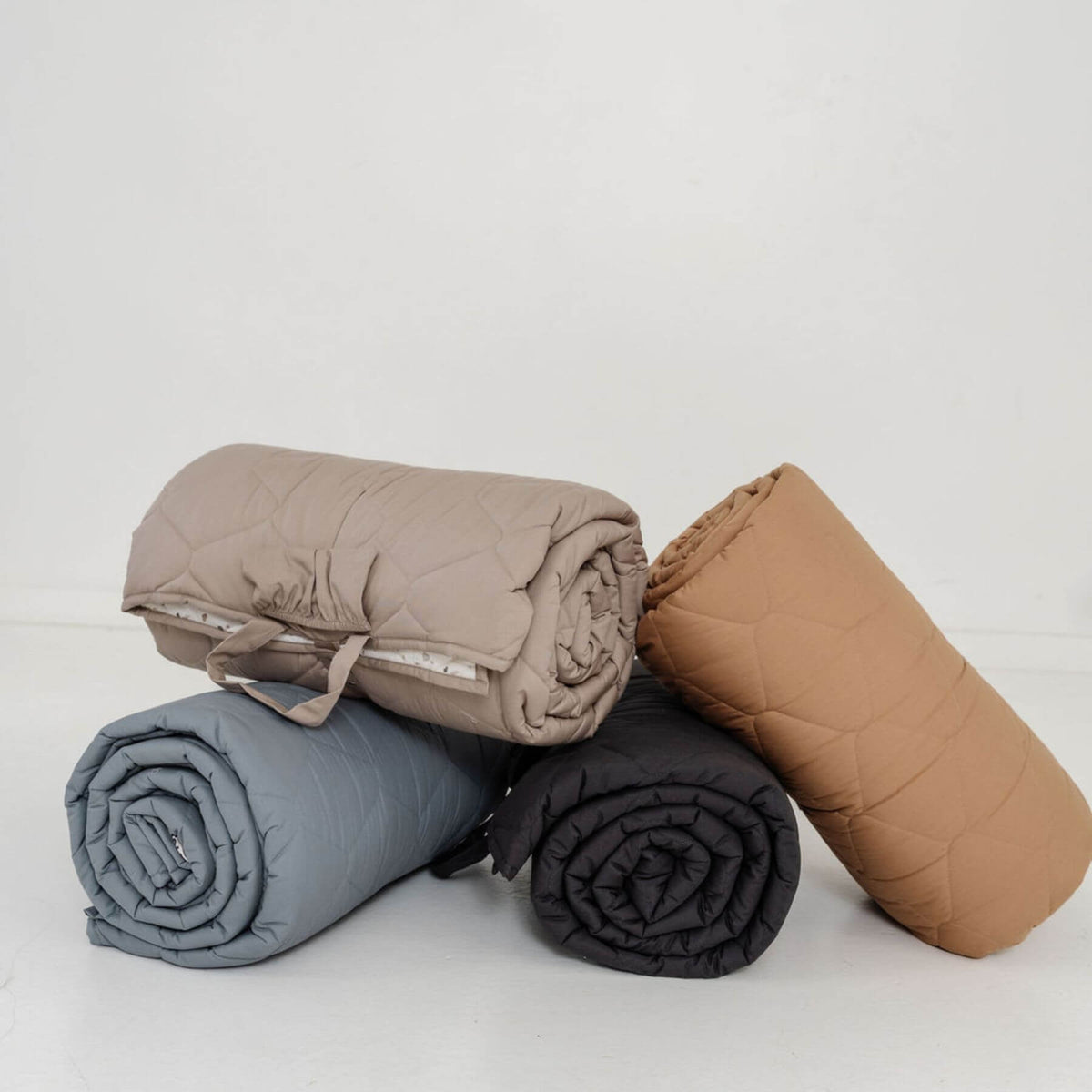 Four rolled-up blankets in brown, gray, and black on a light gray background
