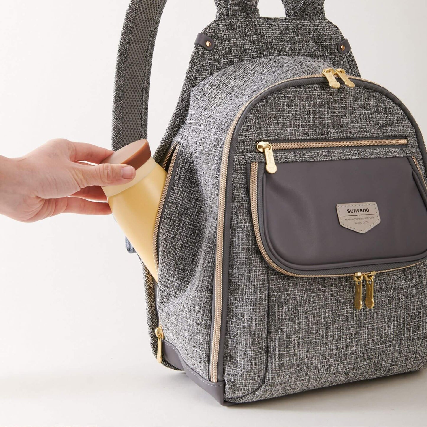 Sunveno Tweed Luxe Diaper Bag Backpack - Gray with a hand holding a yellow item, on a light gray background