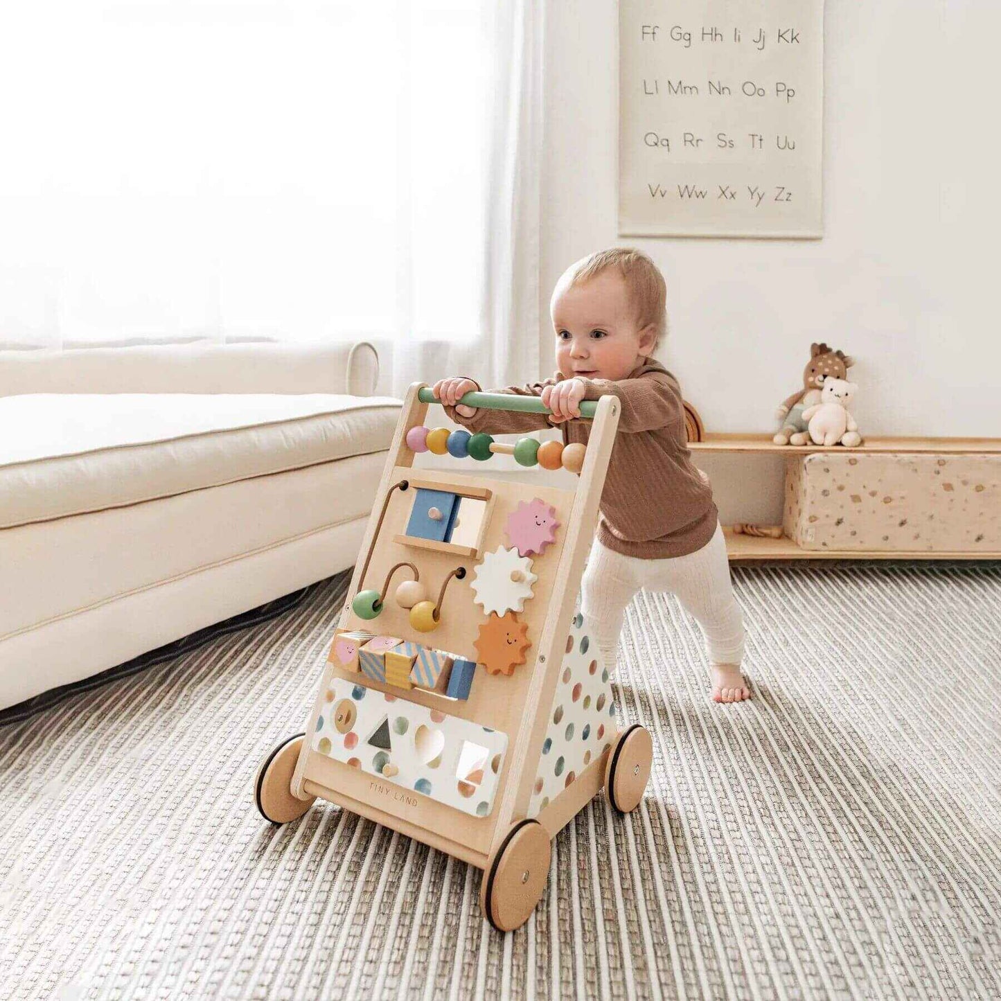 Baby pushing Tiny Land® Versatile Natural Wooden Baby Walker in a room with a white wall and toys.