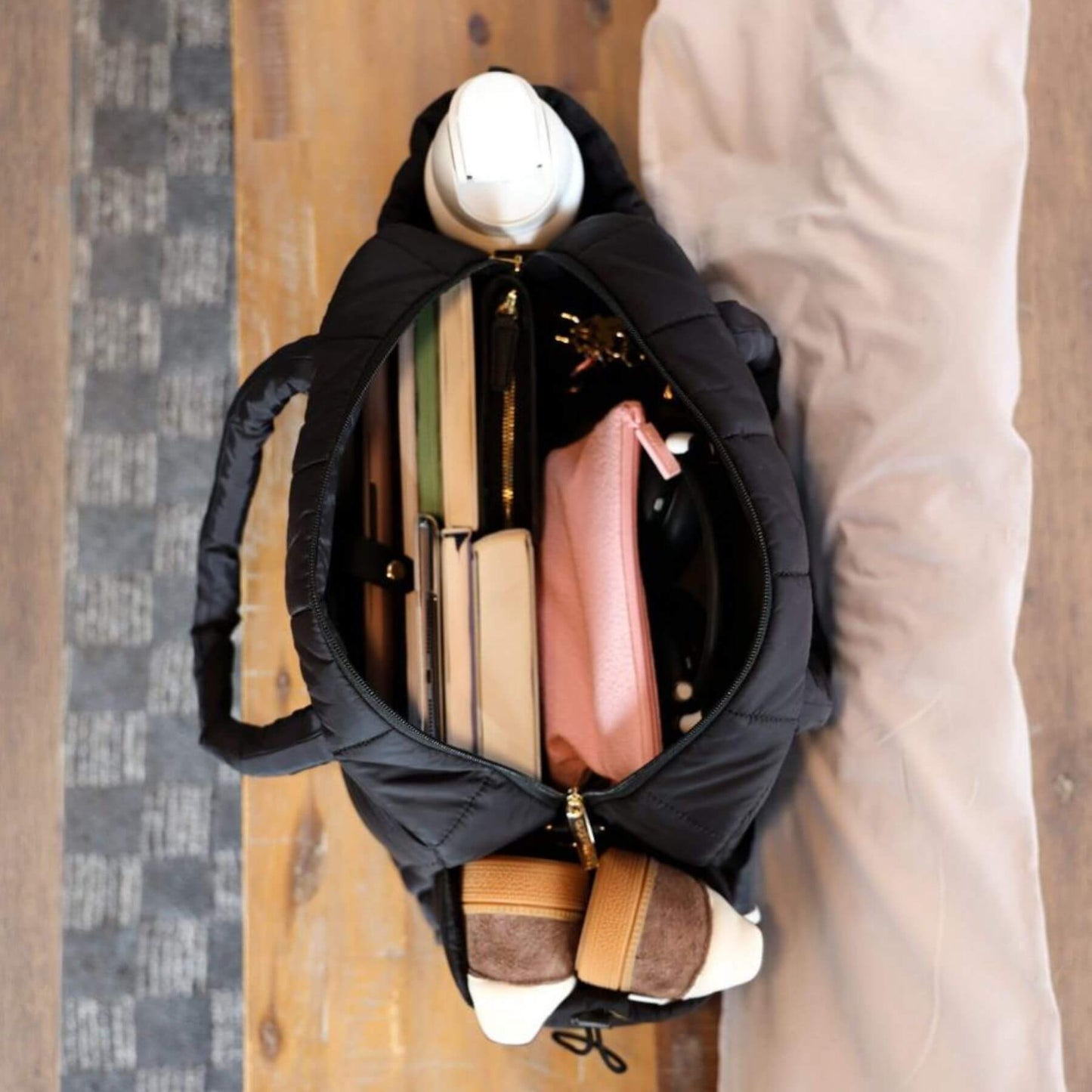 augustnoa the weekender duffle bag black with various items including a pink wallet and a white helmet on a wooden floor.