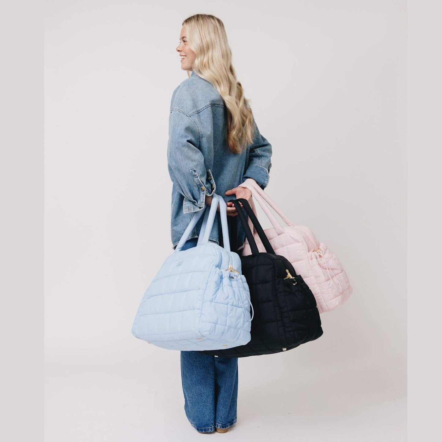 Woman holding three augustnoa the weekender duffle bag in blue, black, and pink.