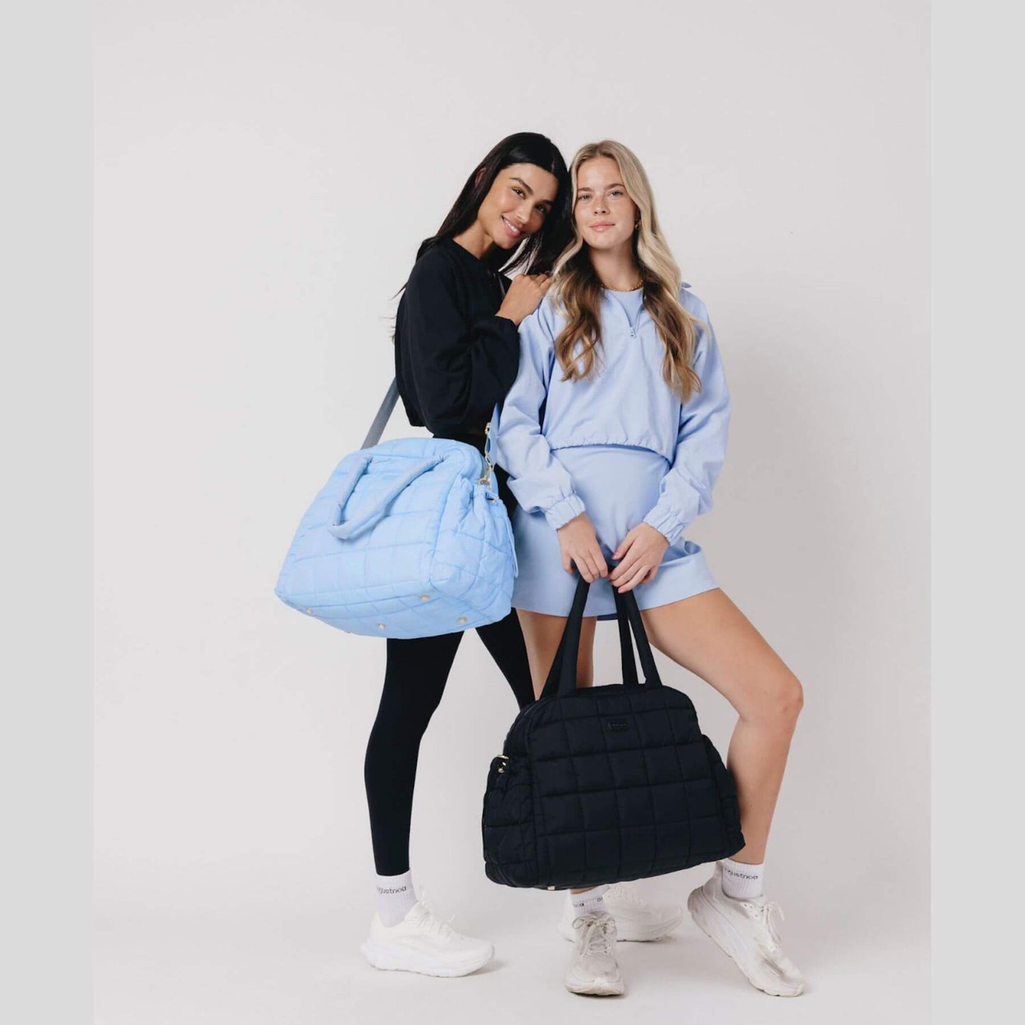 Two women holding a blue and a black augustnoa the weekender duffle bag