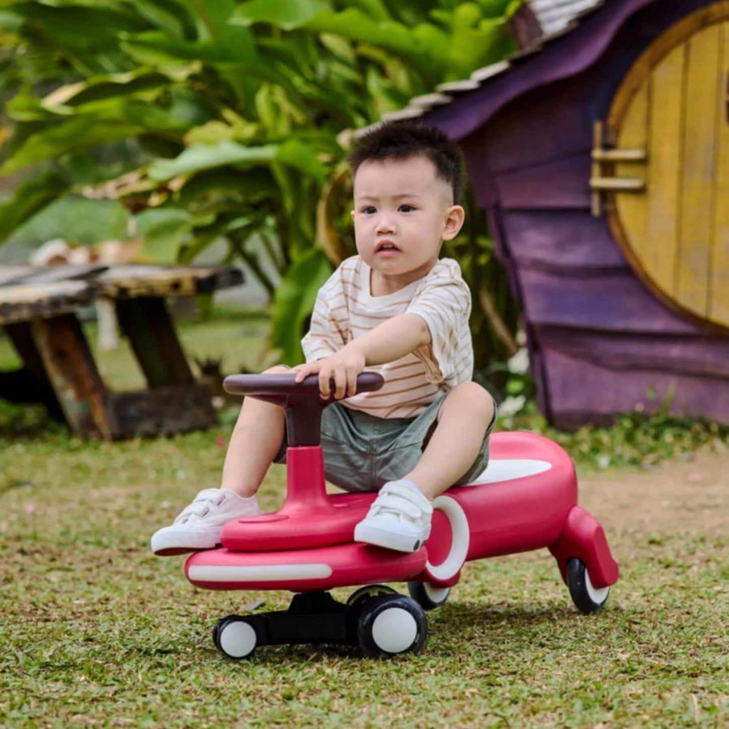 Child sitting on Hape Wiggle & Giggle Car – Red in an outdoor setting with greenery and a small wooden house.