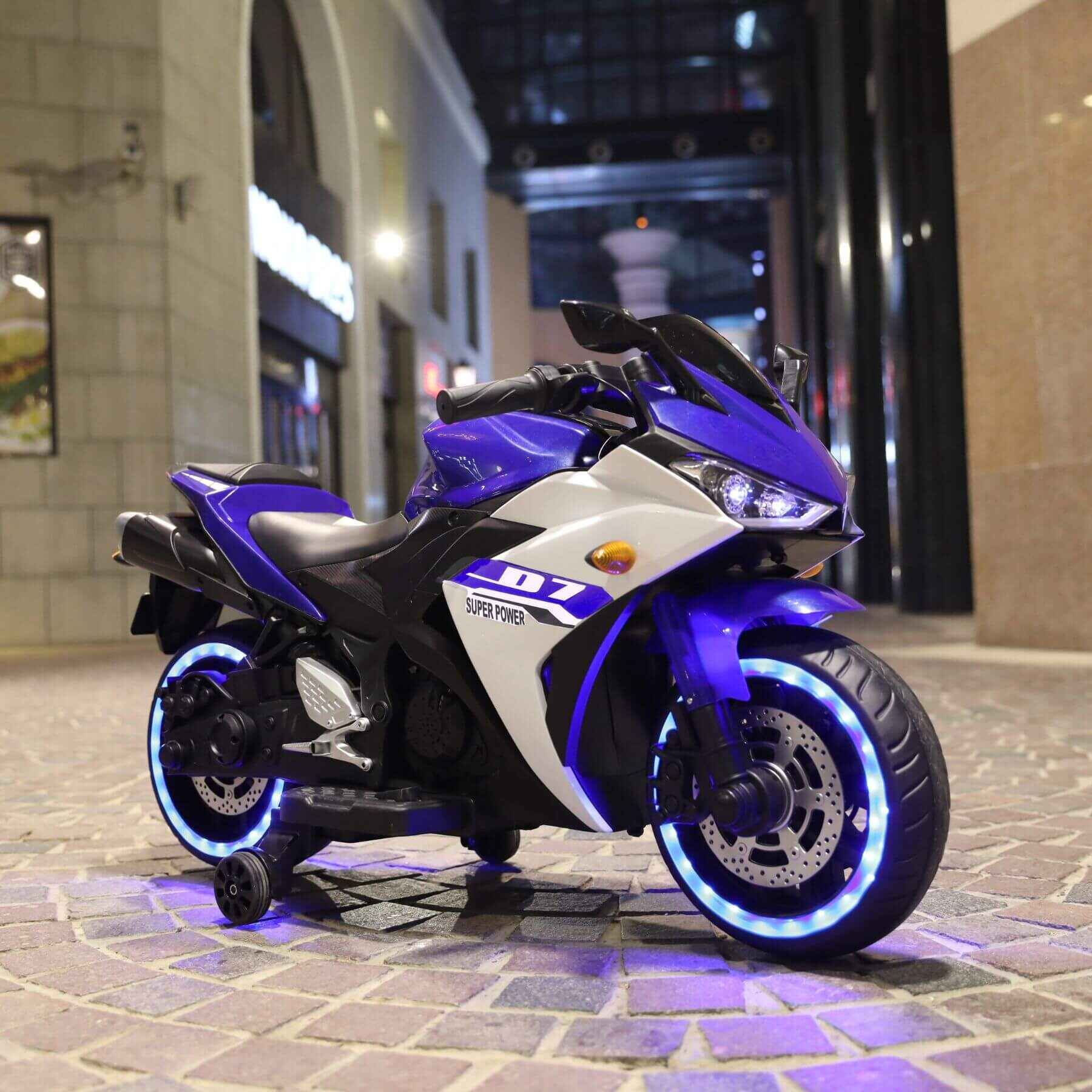 Tamco 12 V Kids Toys Electric Motorcycle with Training Wheels - Blue on a pavement at night.