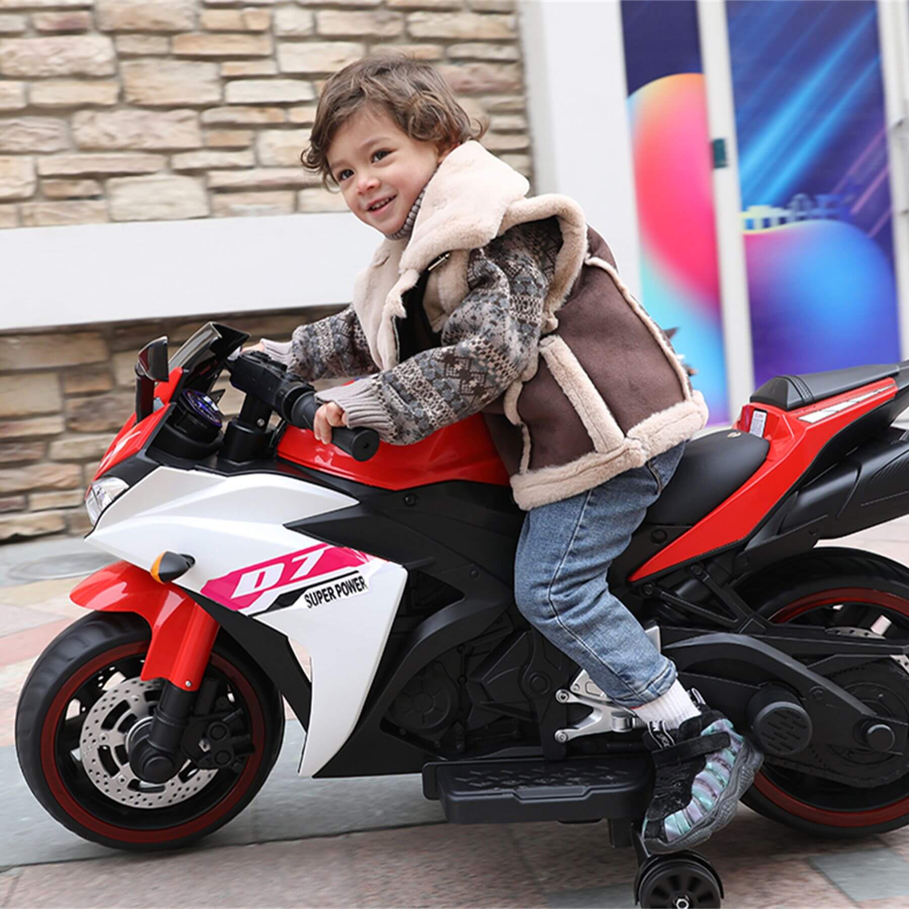 Child sitting on Tamco 12 V Kids Toys Electric Motorcycle with Training Wheels - Red with a colorful background