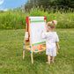 Child drawing on Tiny Land® 3-in-1 Art Easel & Paper Roll Set in a grassy area with trees and flowers in the background