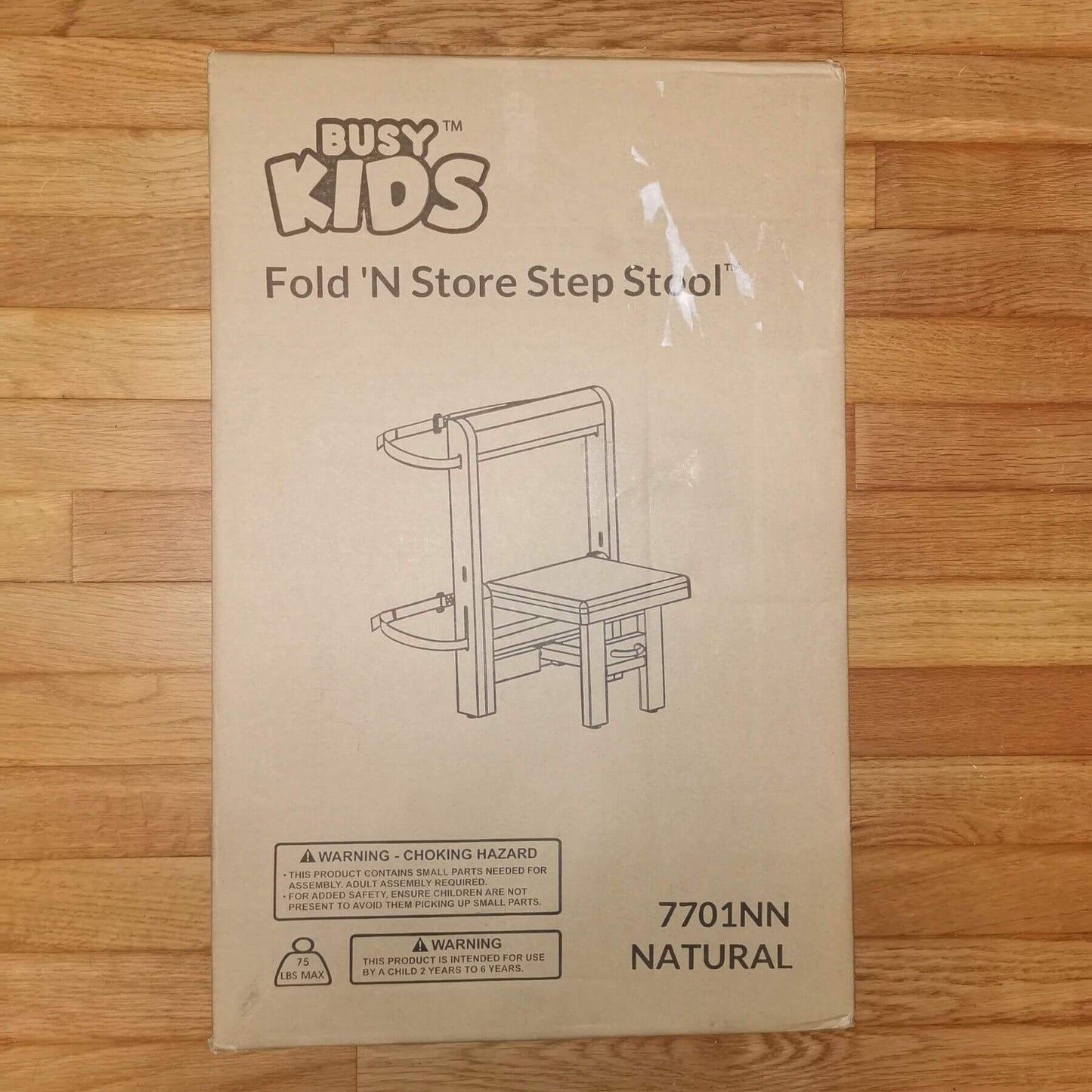 A cardboard product box for a Busy Kids Fold 'N Store Step Stool, shown on a light wood floor.