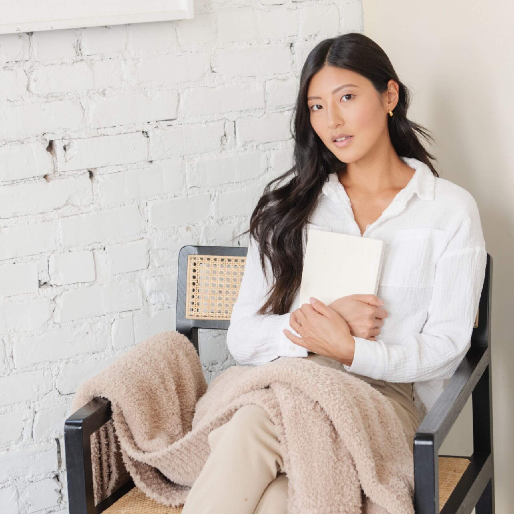 Woman sitting on a chair with BAMBONI® XL Blanket - Birch, holding a book against a white brick wall.
