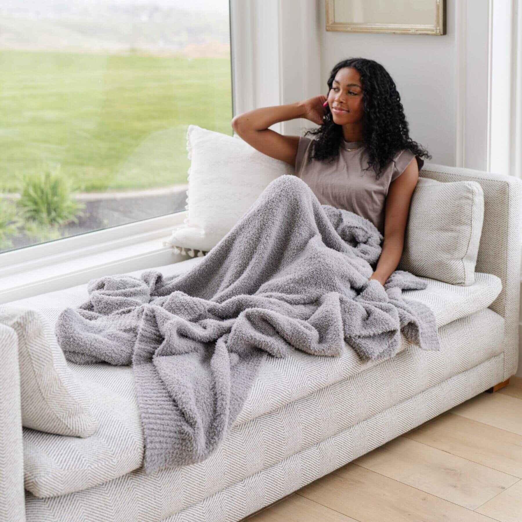 Woman sitting on a couch with BAMBONI® XL Blanket - Gray, in a bright room with large windows.