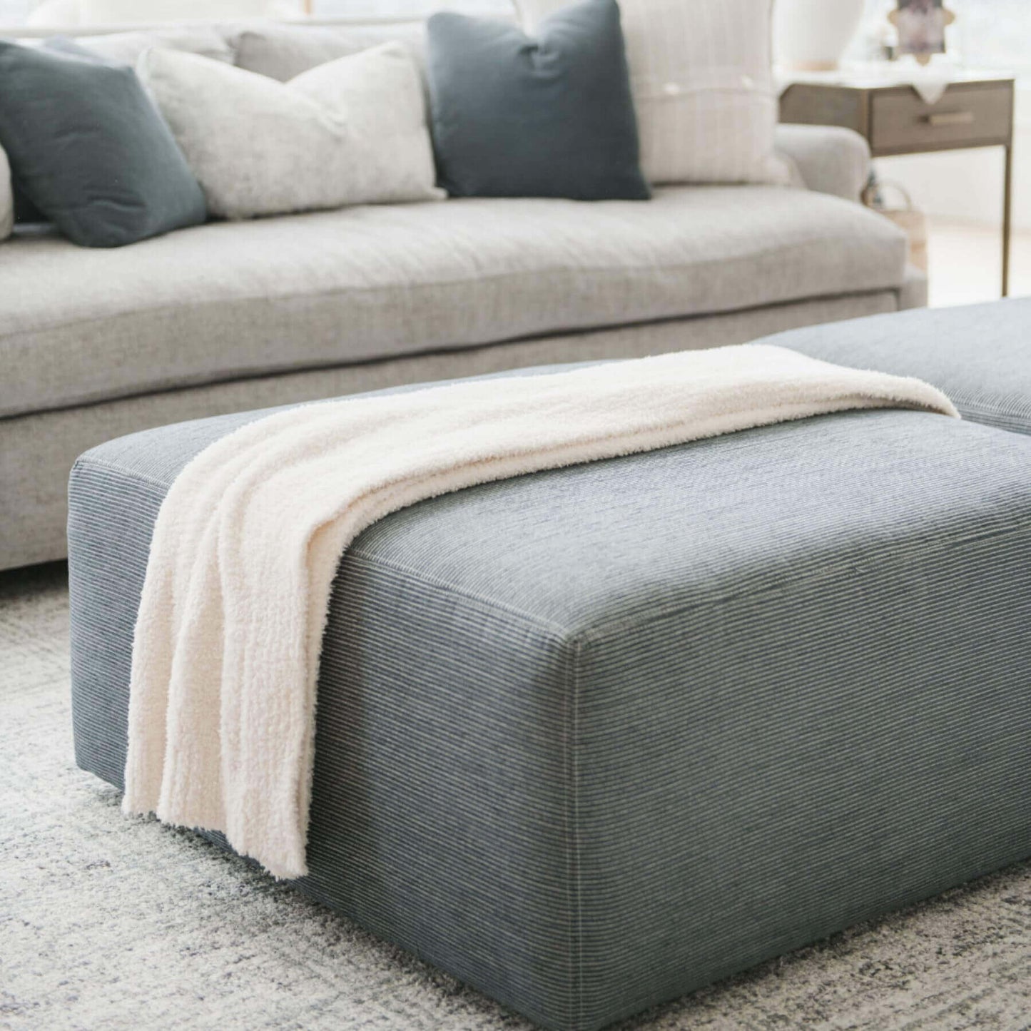 Gray ottoman with BAMBONI® XL Blanket - Ivory draped over it in a living room setting.