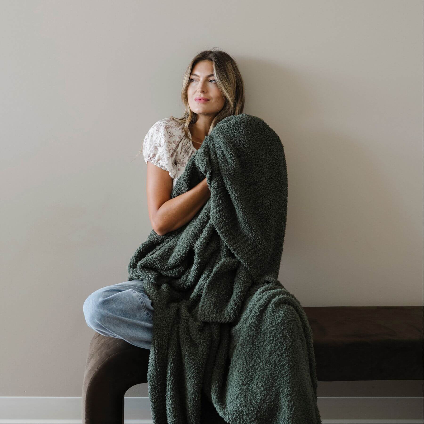 Woman sitting on a chair with BAMBONI® XL Blanket wrapped around her, against a plain wall.