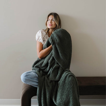 Woman sitting on a chair with BAMBONI® XL Blanket wrapped around her, against a plain wall.