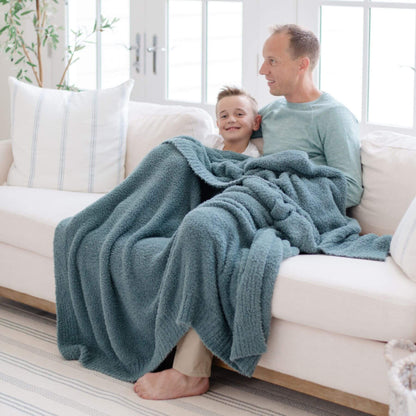 Man and child sitting on a couch under BAMBONI® XL Blanket - Spruce in a bright living room.