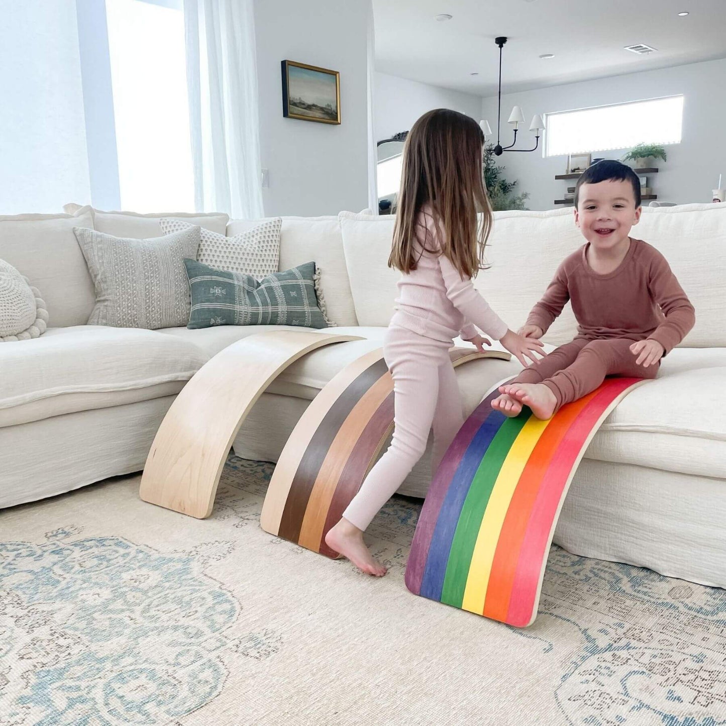 Two children playing on a colorful rainbow-shaped toy in a living room.