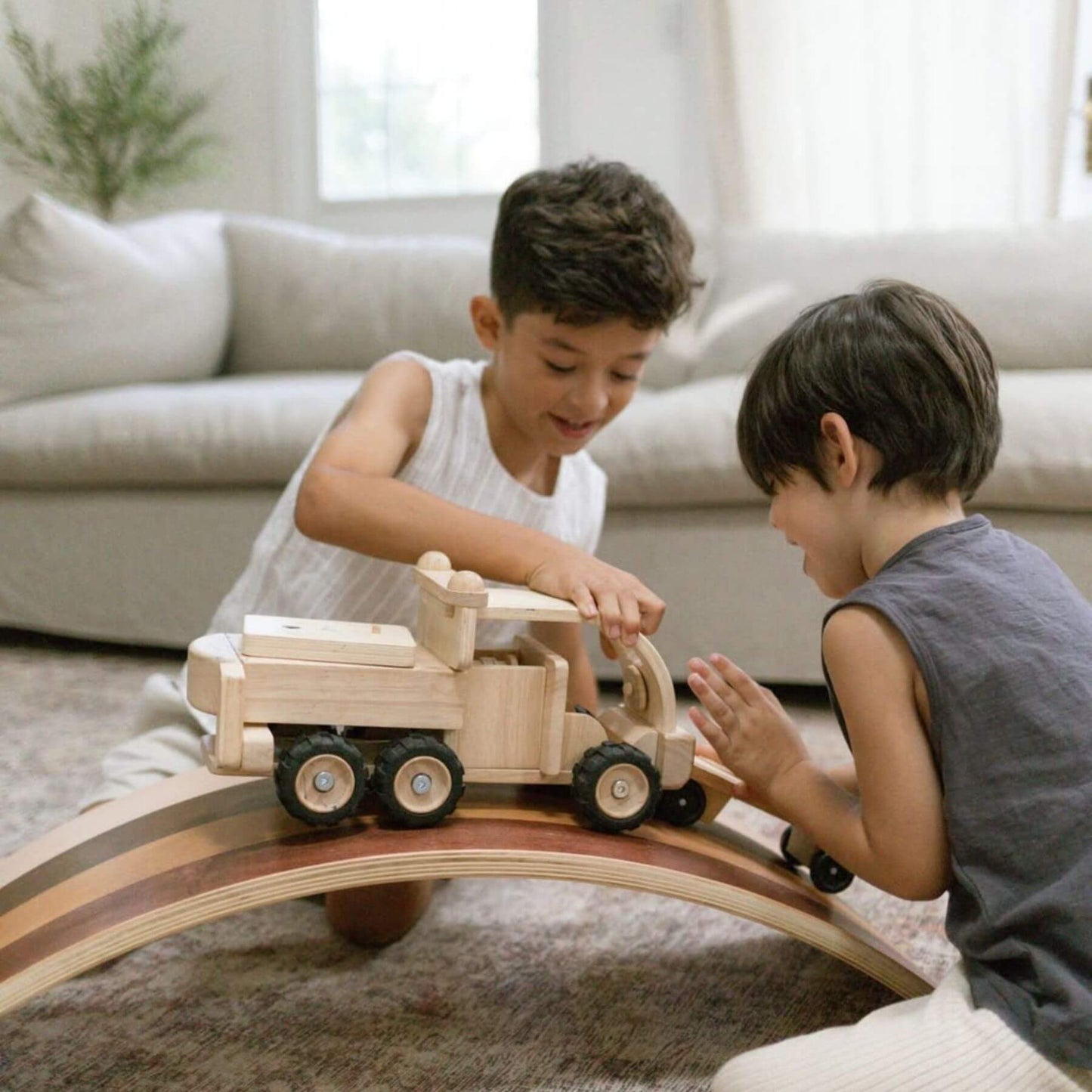 Two children playing with a wooden toy truck on a curved track in a living room.