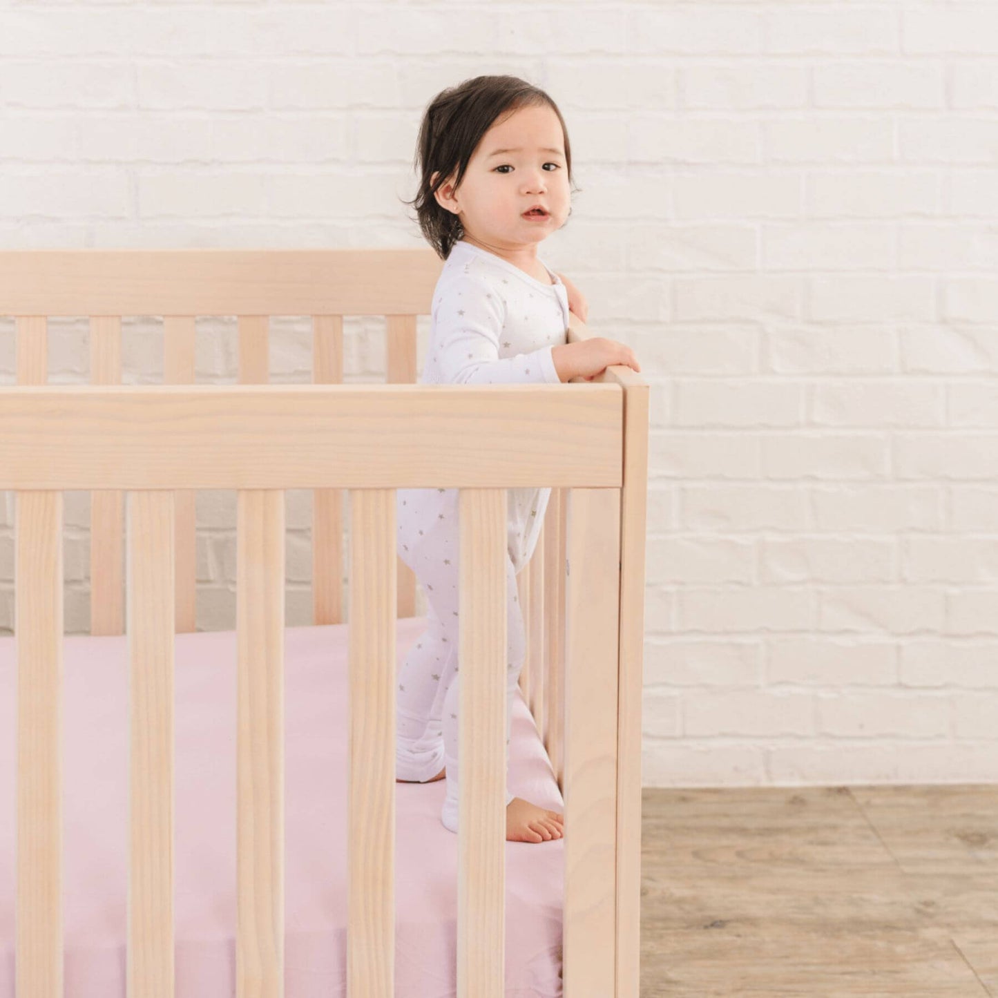 Child standing in a crib with a light wooden frame and pink bedding against a white brick wall.