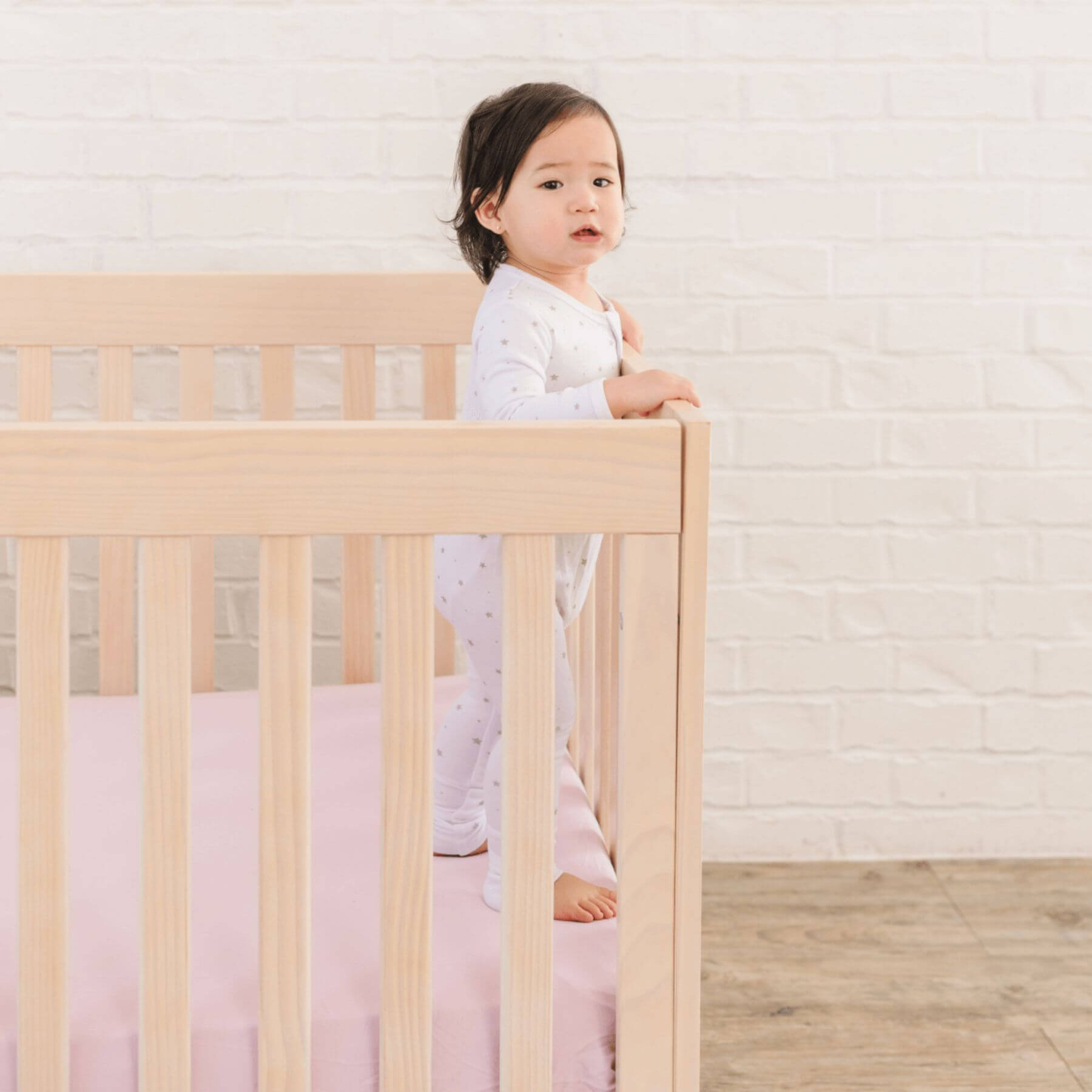 Child standing in a crib with a light wooden frame and pink bedding against a white brick wall.