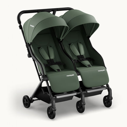 A double stroller in sage green fabric with a black frame and wheels, shown from a three-quarters angle on a light off-white background. The brand name 'monpush' is visible on the side of the stroller seats.