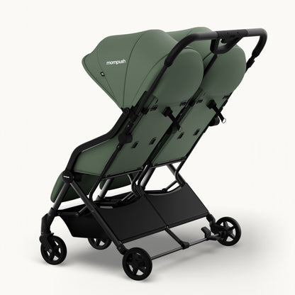 A compact double stroller in sage green fabric with a black frame and wheels, shown from a three-quarters angle on a light gray background. The brand name 'mompush' is visible on the canopy.