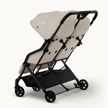 A compact double stroller in a light beige fabric with a black frame, shown from a three-quarters angle on a clean white background. The brand name 'monpush' is visible on the canopy.