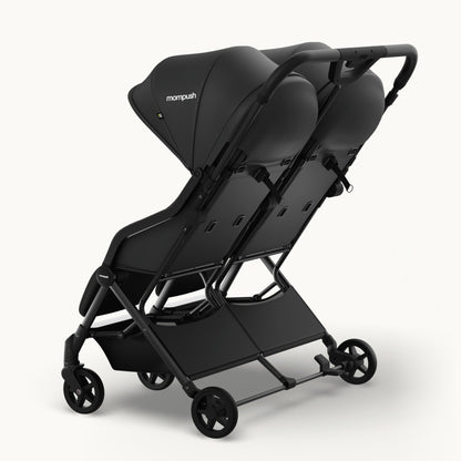 A black Mompush double stroller with a matching canopy, shown from a three-quarters angle on a light gray studio background.