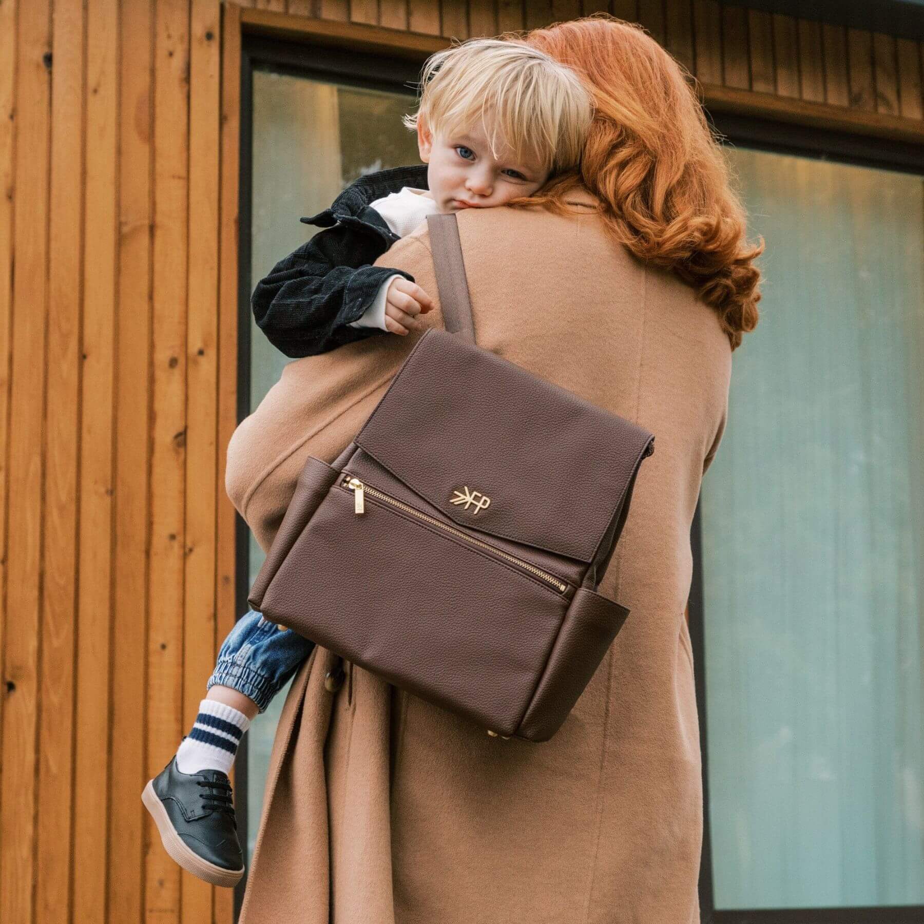 Woman carrying a child on her back with Freshly Picked Espresso Mini Classic Diaper Bag II, standing in front of a wooden building.
