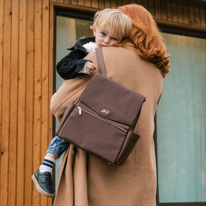 Woman carrying a child on her back with Freshly Picked Espresso Mini Classic Diaper Bag II, standing in front of a wooden building.