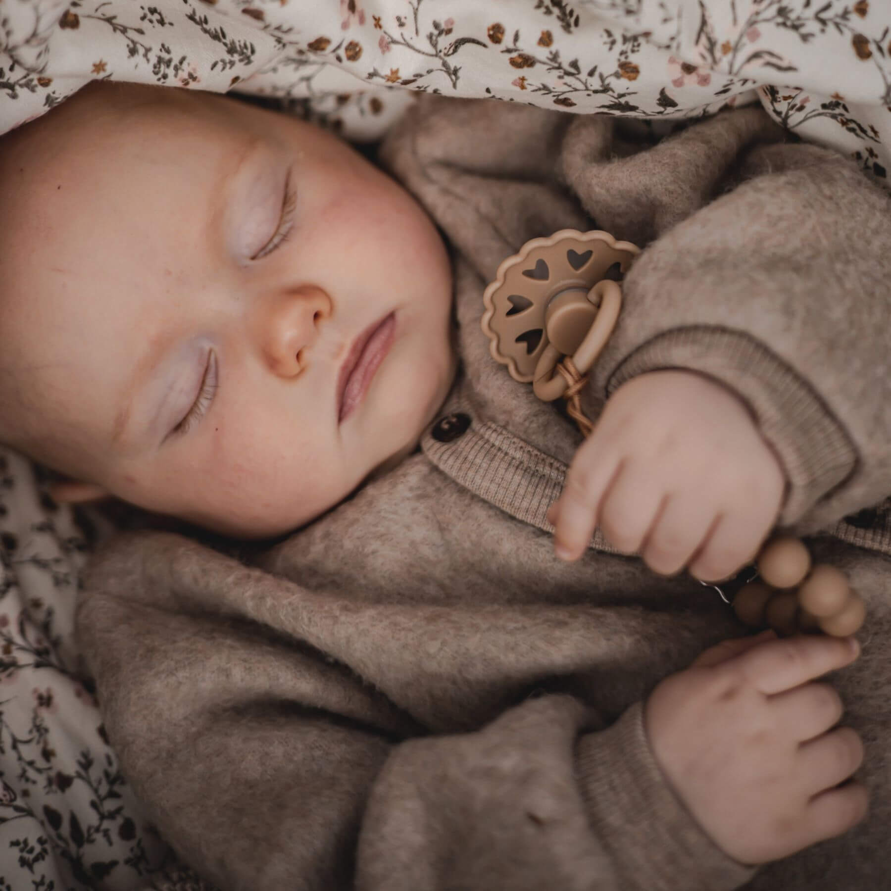 Baby sleeping in a floral crib with a brown outfit and Mushie FRIGG Silicone Pacifier