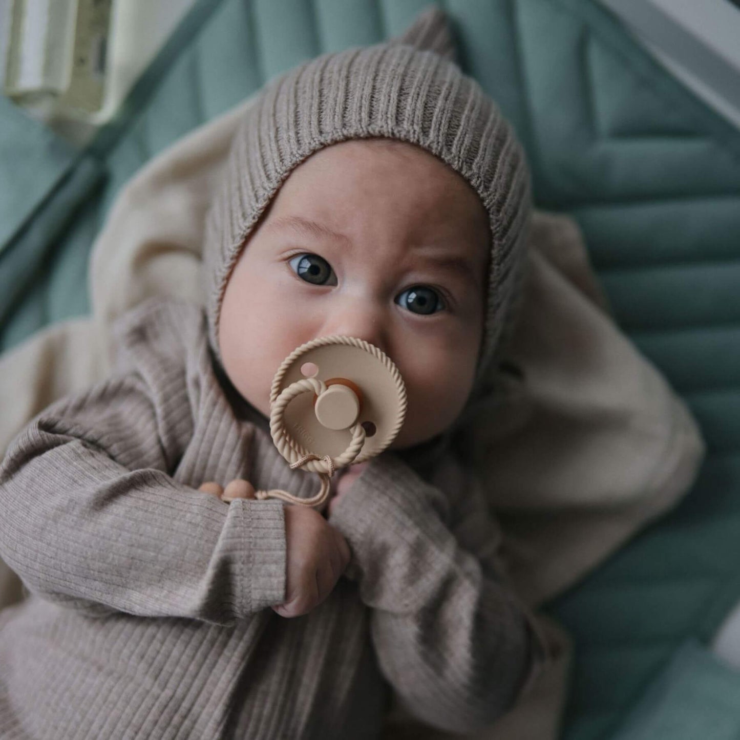 Baby in a beige outfit holding Mushie FRIGG Silicone Pacifier, sitting on a teal cushion.