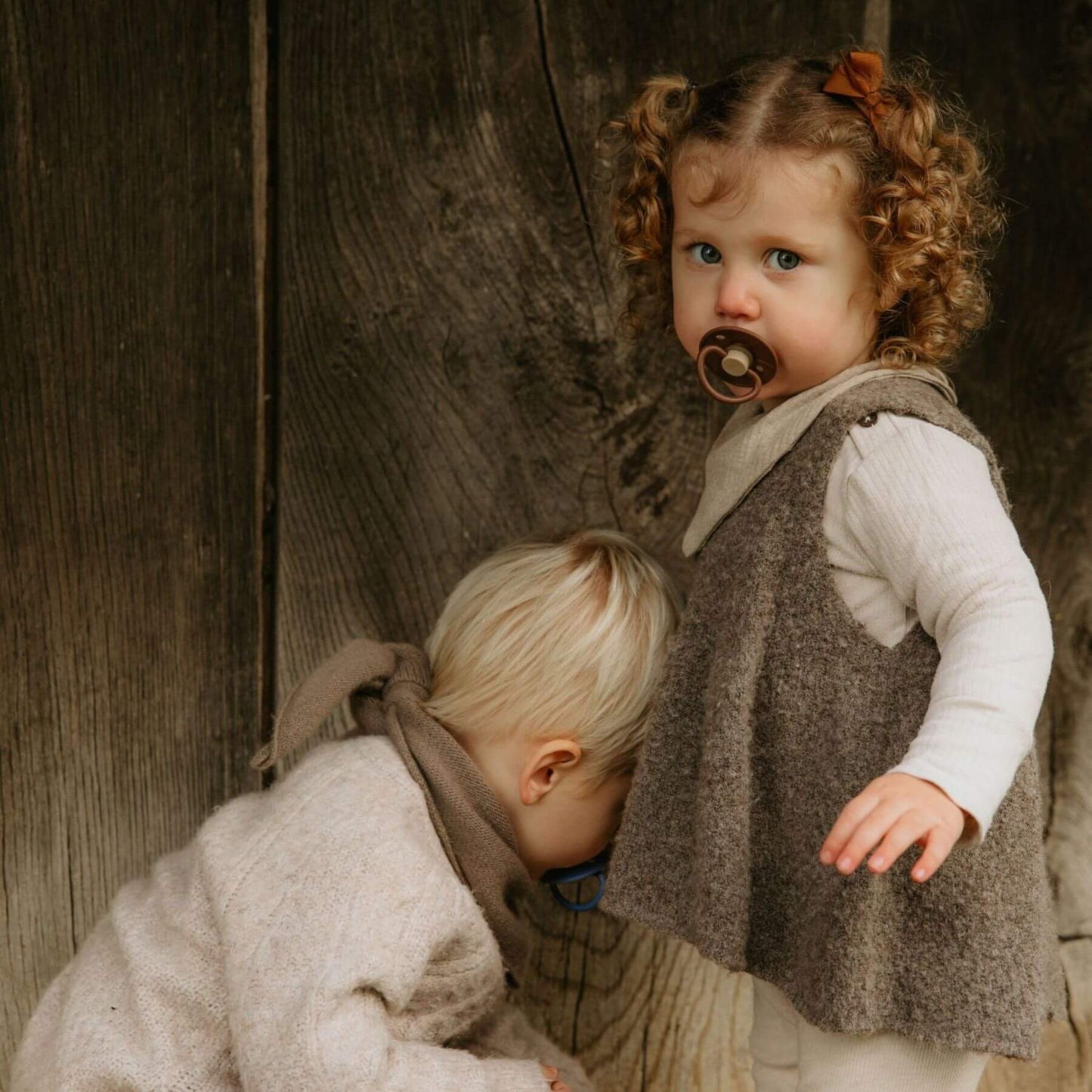 Two children holding Mushie FRIGG Silicone Pacifier, in matching outfits standing against a wooden wall.