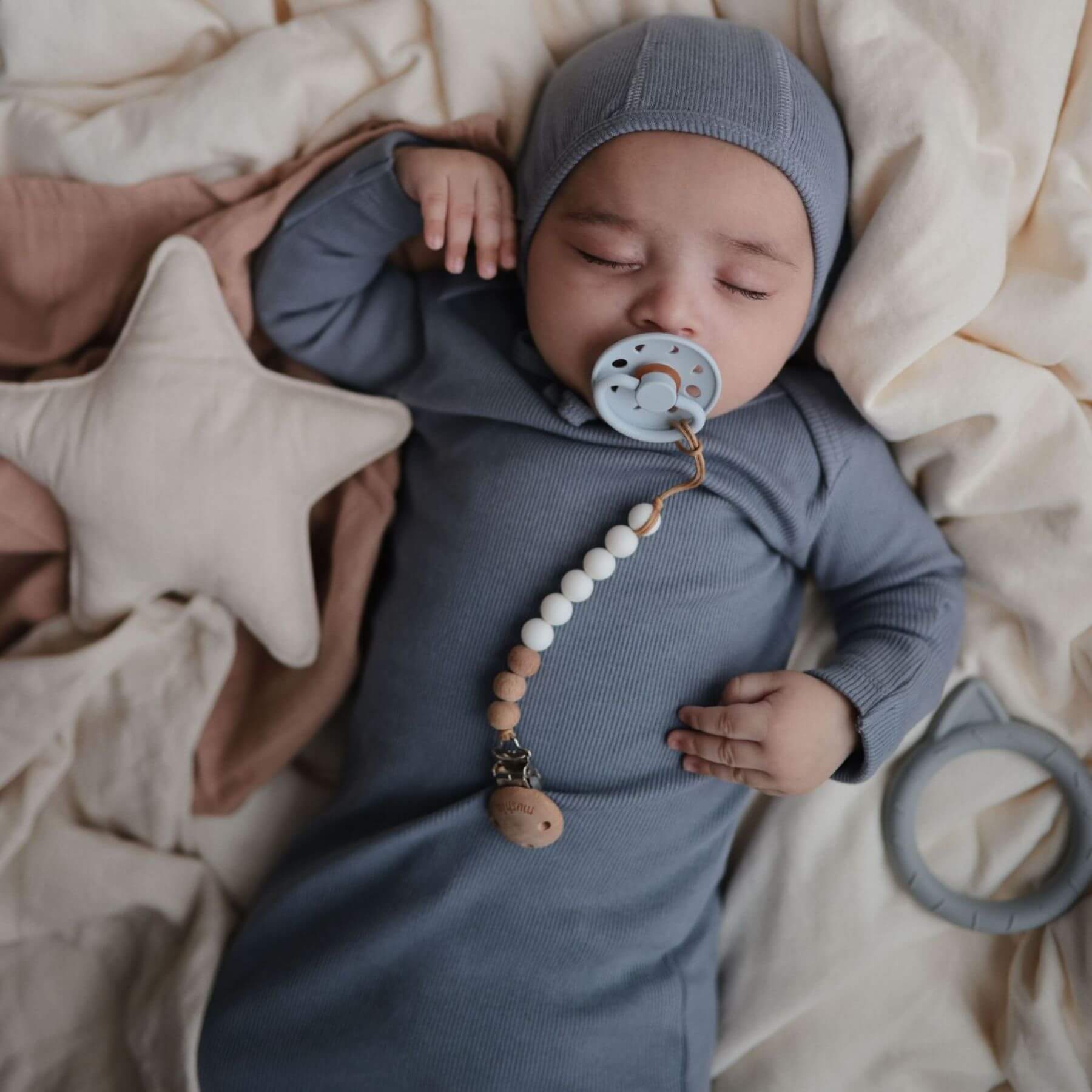 Baby in a blue outfit with Mushie FRIGG Silicone Pacifier and teething ring, surrounded by soft textures and toys.