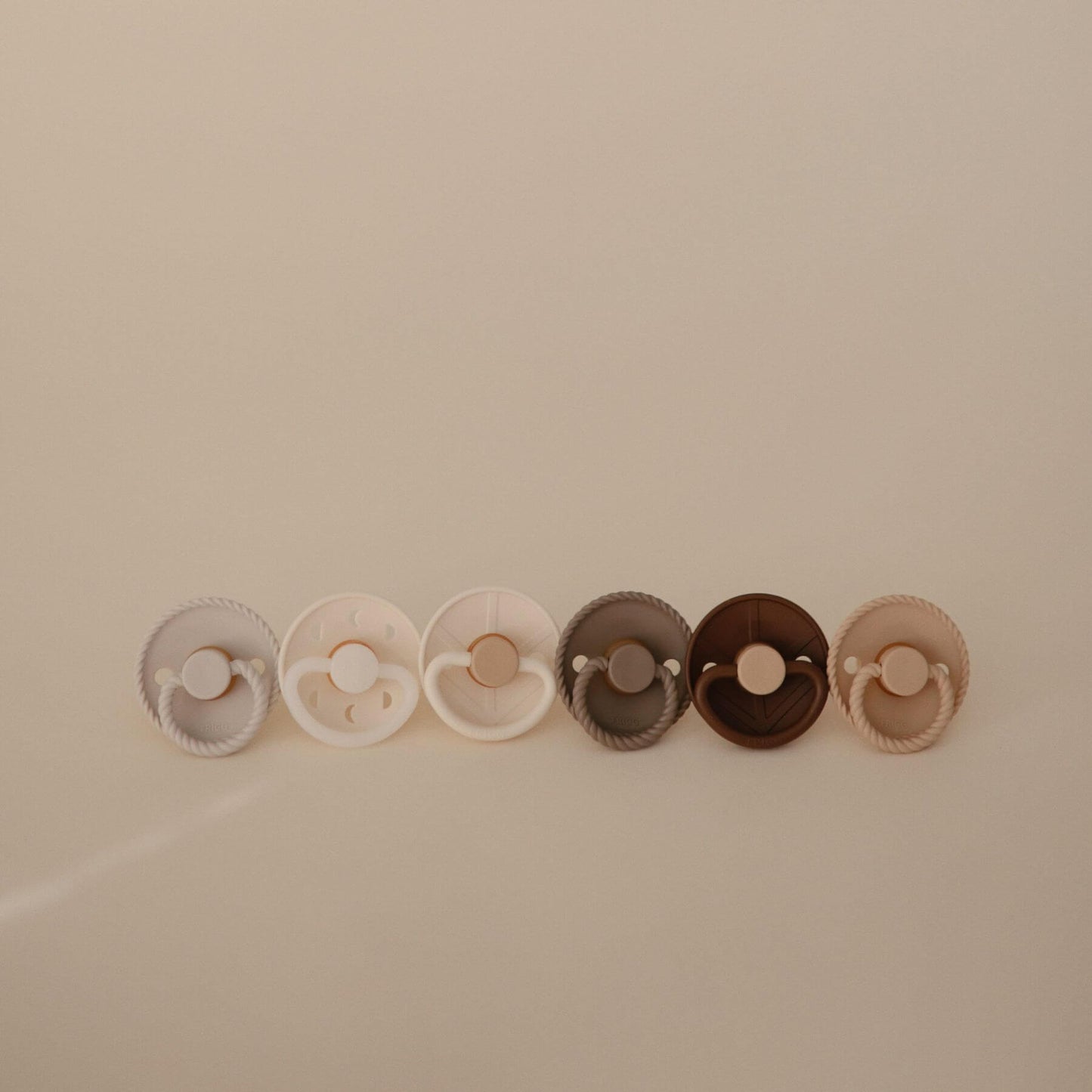 Set of five Mushie FRIGG Silicone Pacifier in various colors arranged in a row on a beige background
