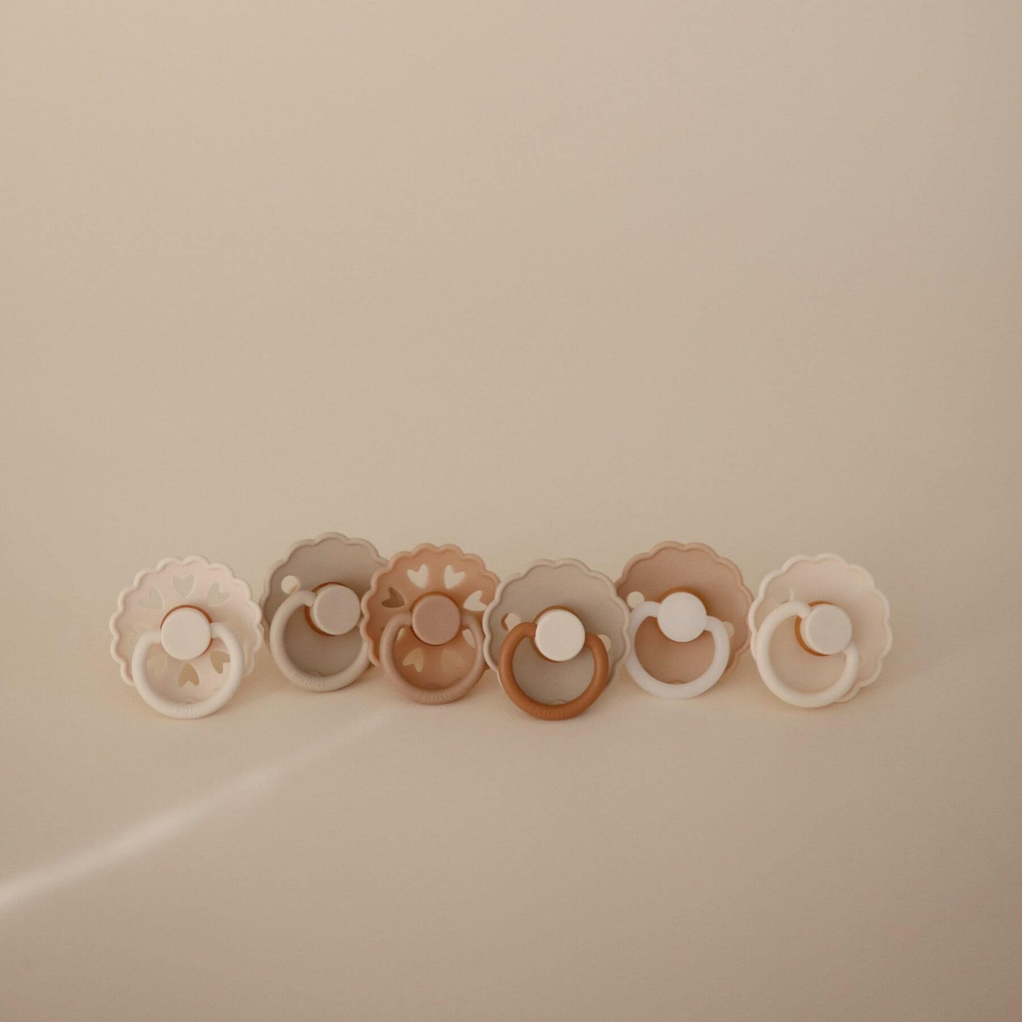 Set of six Mushie FRIGG Silicone Pacifiers in various colors on a beige background