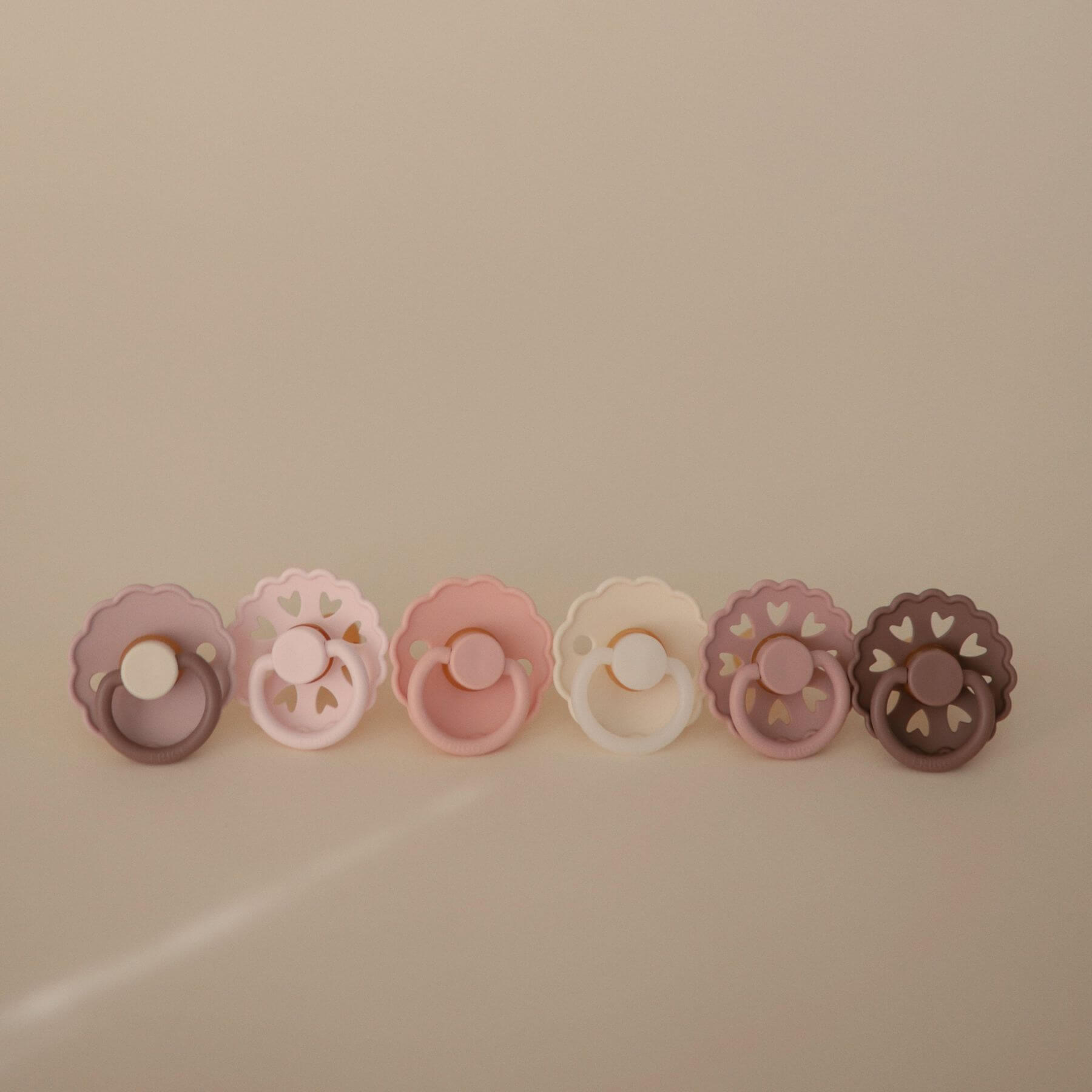 Set of six Mushie FRIGG Silicone Pacifiers in various colors on a beige background