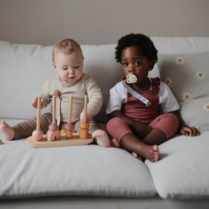 Two children sitting on a couch with a wooden toy and Mushie FRIGG Silicone Pacifier