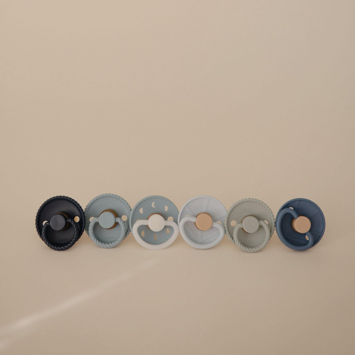 Set of five Mushie FRIGG Silicone Pacifiers in various colors on a beige background