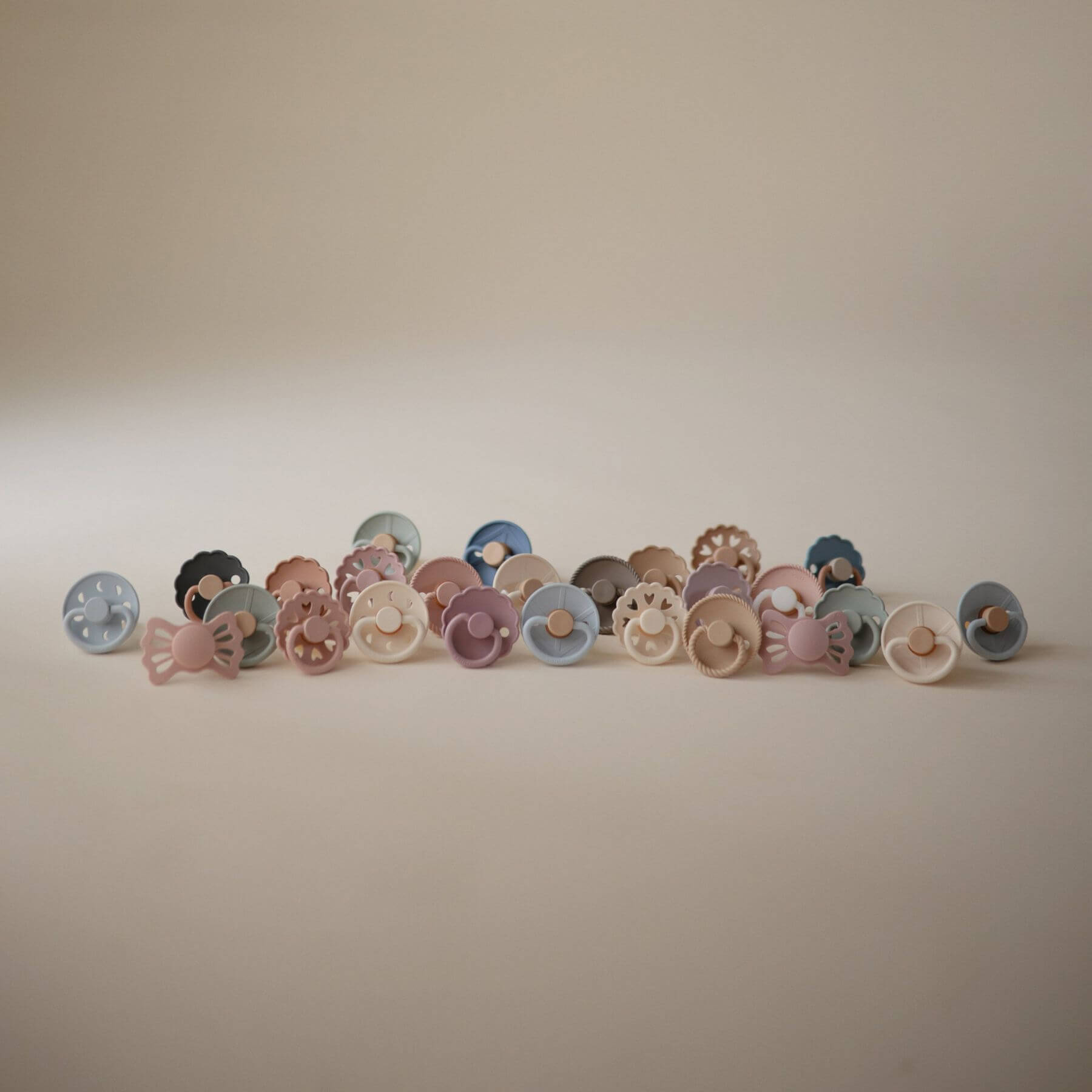 Row of colorful Mushie FRIGG Silicone Pacifiers on a beige background