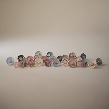 Row of colorful Mushie FRIGG Silicone Pacifiers on a beige background