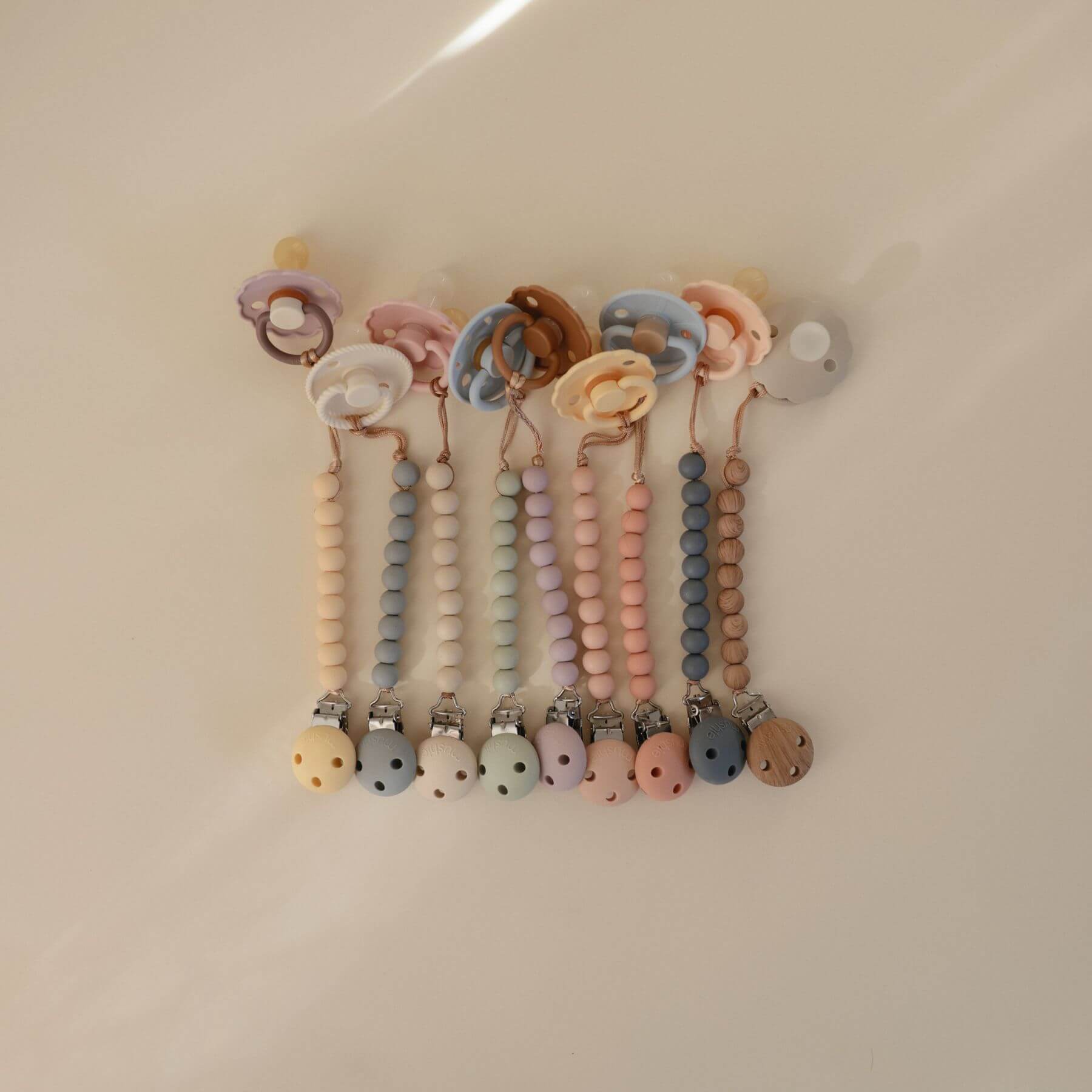 Set of colorful baby Mushie FRIGG Silicone Pacifier clips with beads on a beige background