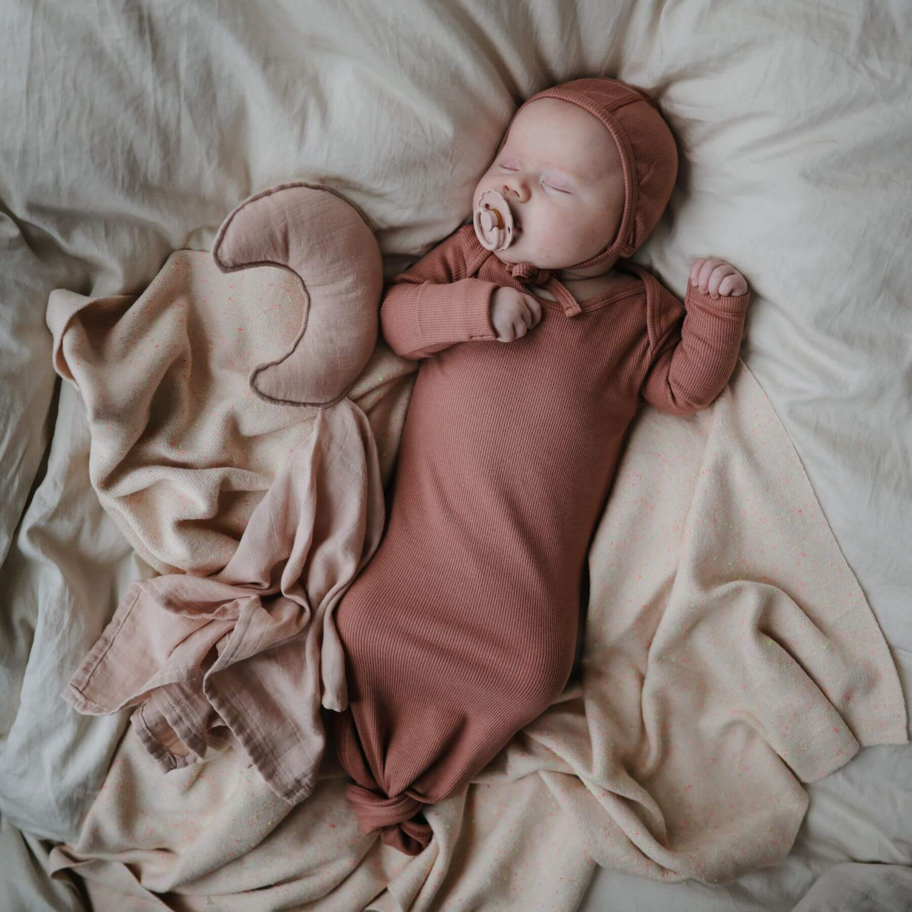 Newborn baby in a pink outfit and hat lying on a soft beige blanket, with Mushie FRIGG Silicone Pacifier