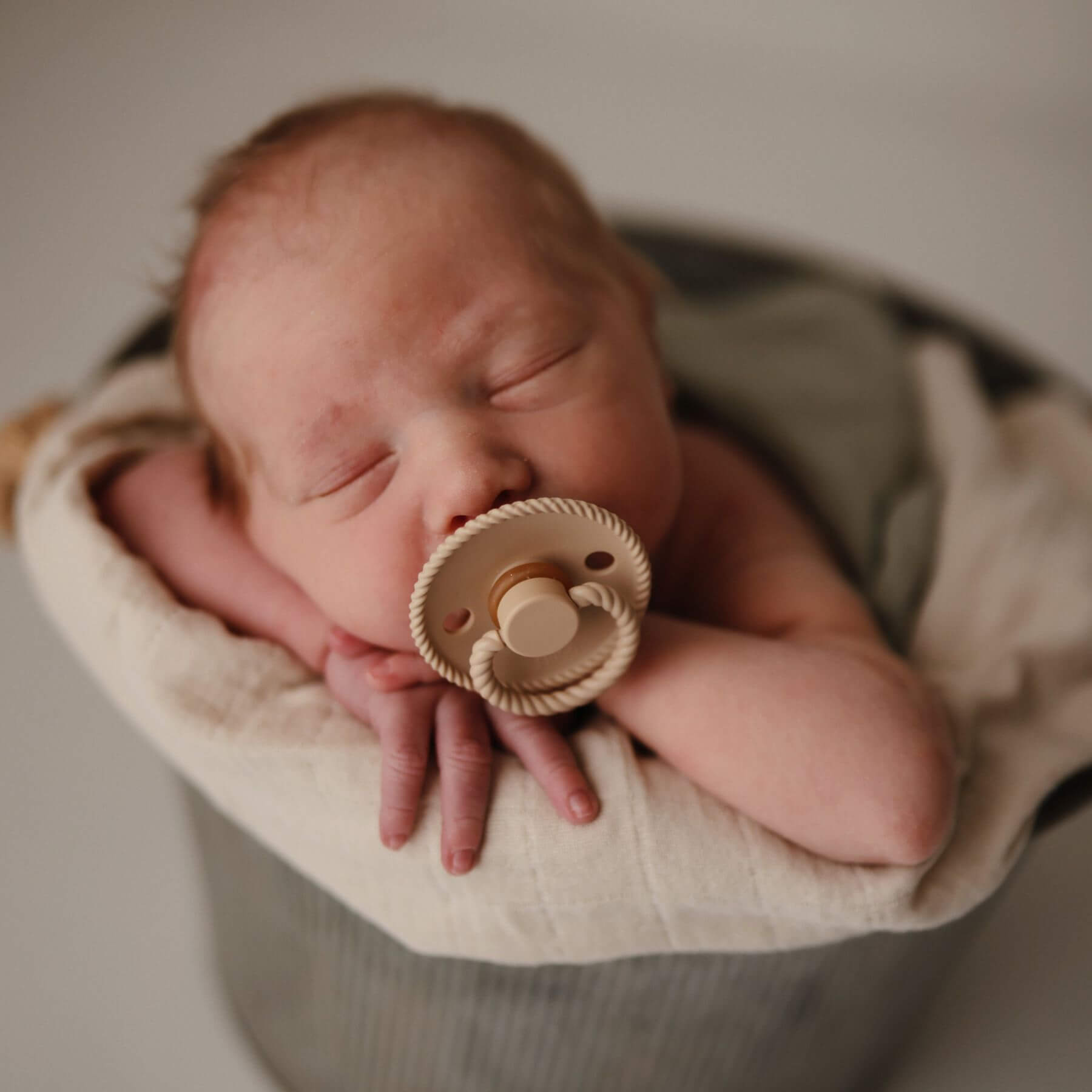 Newborn baby holding Mushie FRIGG Silicone Pacifier wrapped in soft fabric