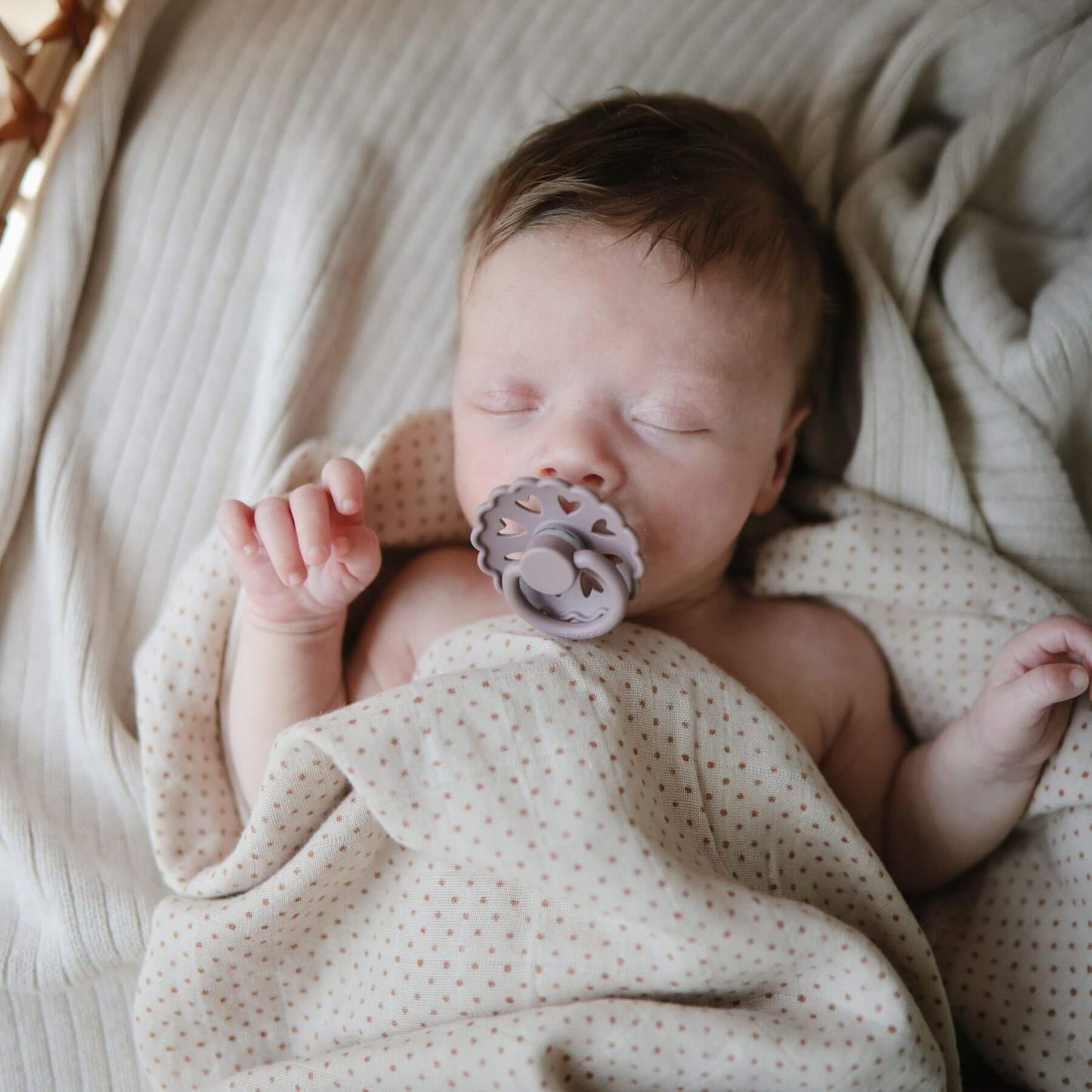 Newborn baby sleeping with Mushie FRIGG Silicone Pacifier, wrapped in a beige blanket.