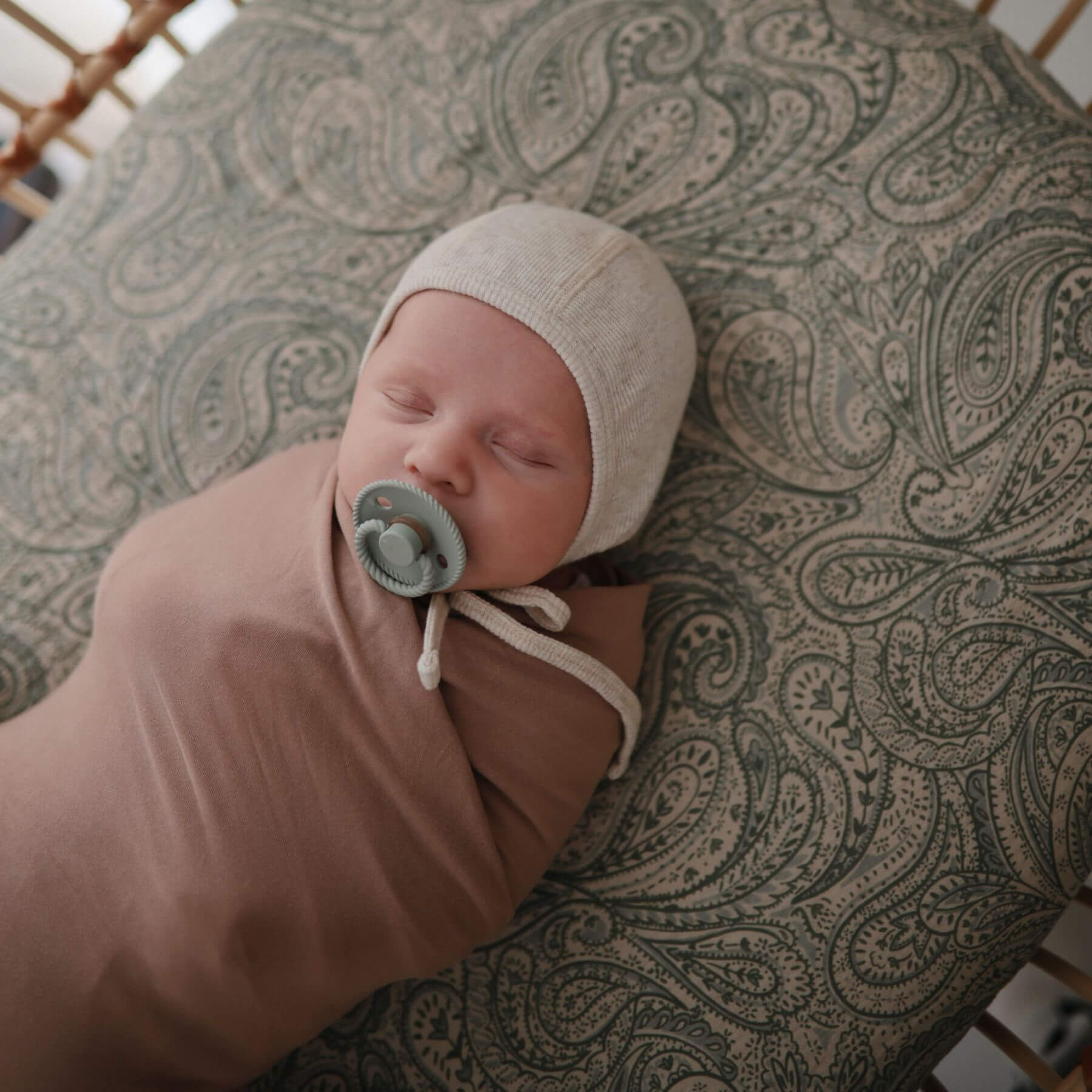 Newborn baby sleeping with Mushie FRIGG Silicone Pacifier, wearing a beige bonnet, on a patterned surface.