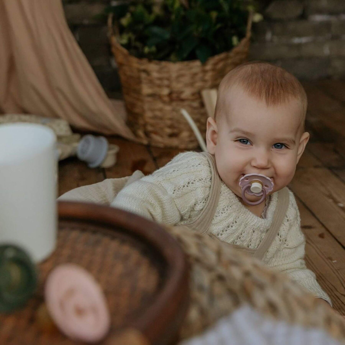Baby in a knitted outfit with Mushie FRIGG Silicone Pacifier, surrounded by rustic elements.